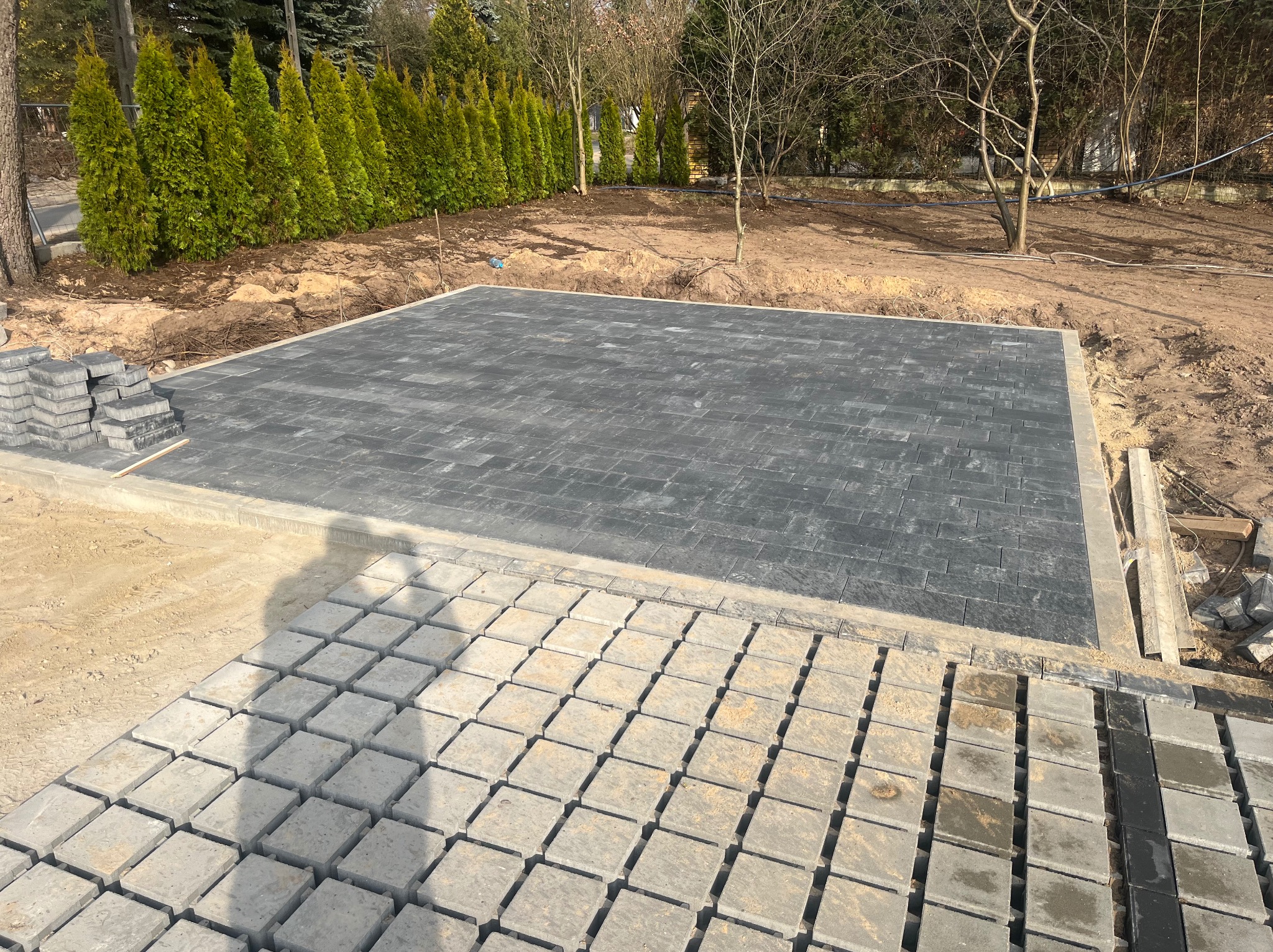 Częściowo ułożona, szara kostka brukowa na przygotowanym podłożu. W tle rząd zielonych drzew i piasek. Widoczne zapasy kostki brukowej w dwóch odcieniach szarości.
