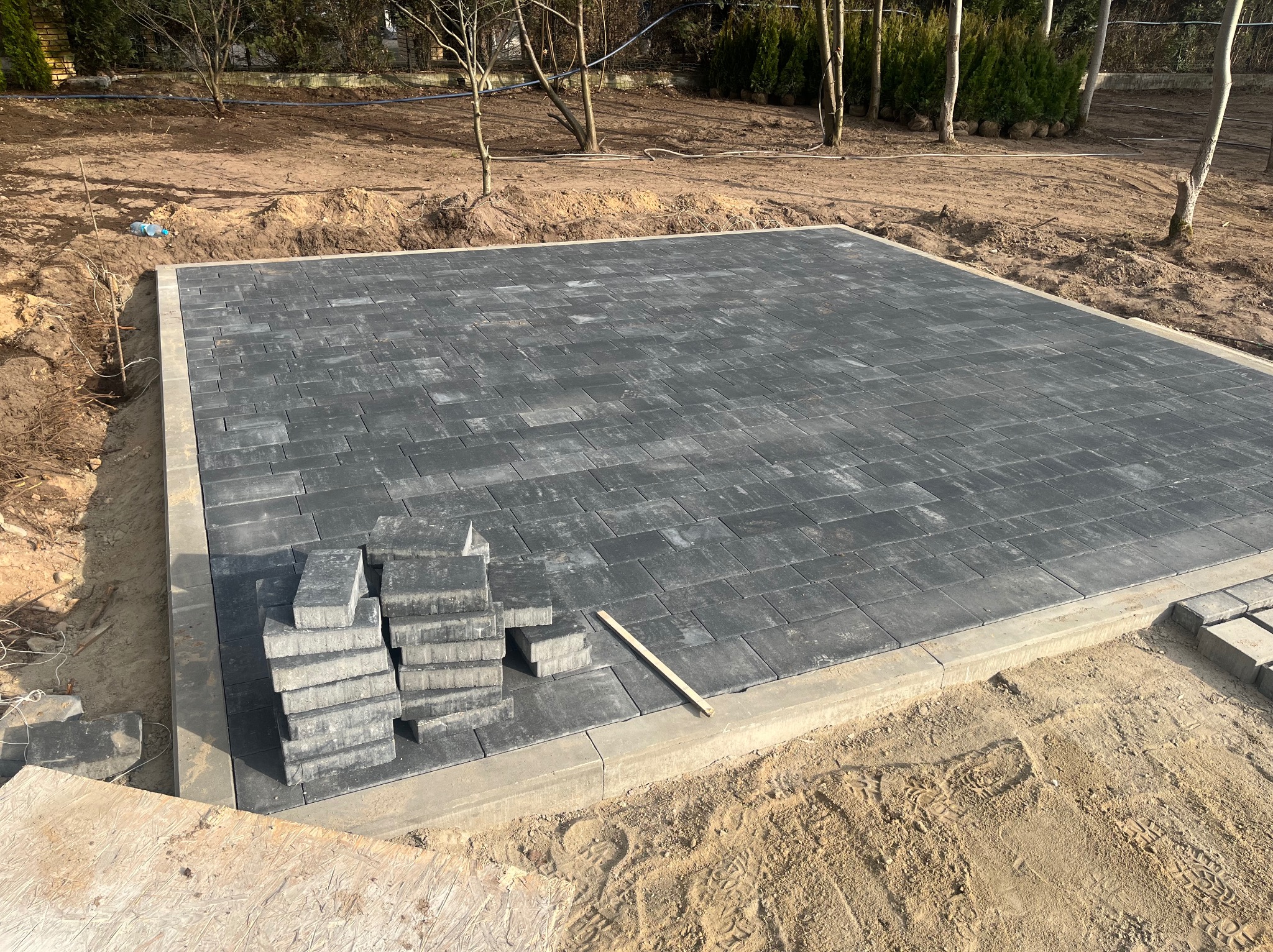 Częściowo ułożona, szara kostka brukowa na piasku, obramowana betonowym krawężnikiem. Stos kostki i drewniana listwa obok. Tło: ziemia i drzewa.