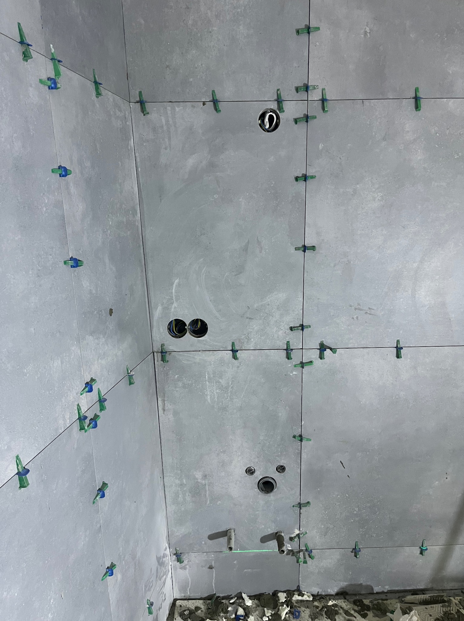 Szare płytki ścienne w trakcie montażu, z użyciem klipsów poziomujących i krzyżyków dystansowych. Widoczne otwory na instalacje. Ujęcie z dołu, surowy stan.