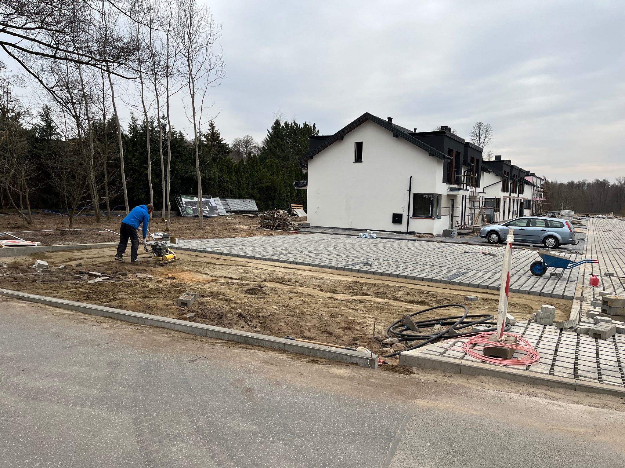 Układanie kostki brukowej na posesji szeregowej zabudowy. Pracownik zagęszcza podłoże ubijarką. Częściowo ułożona kostka, krawężniki, narzędzia i materiały budowlane.