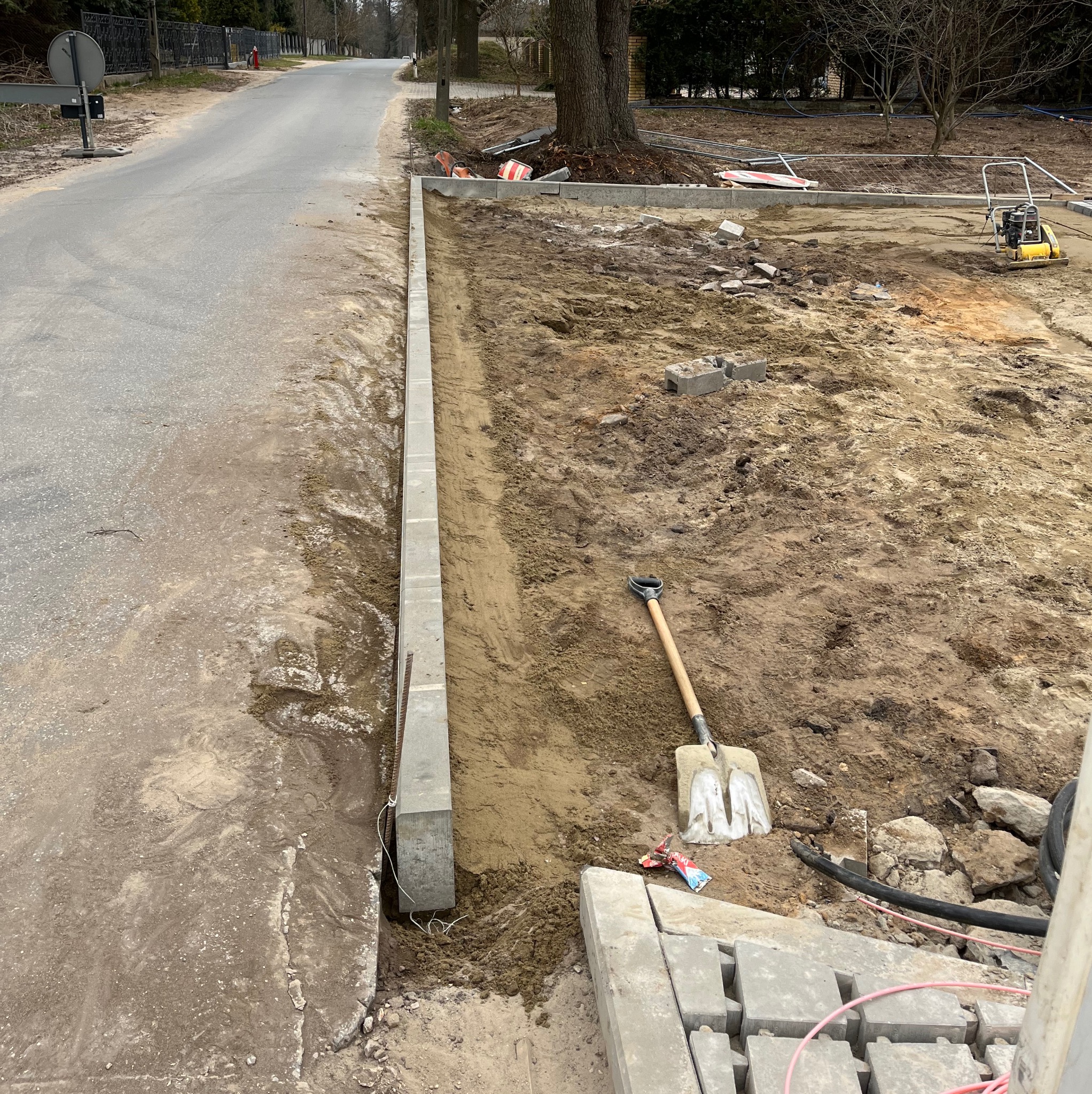 Montaż obrzeży chodnikowych z betonu. Widoczny wykop, ułożone krawężniki, łopata i zagęszczarka w tle. Prace brukarskie w toku, przygotowanie podłoża.