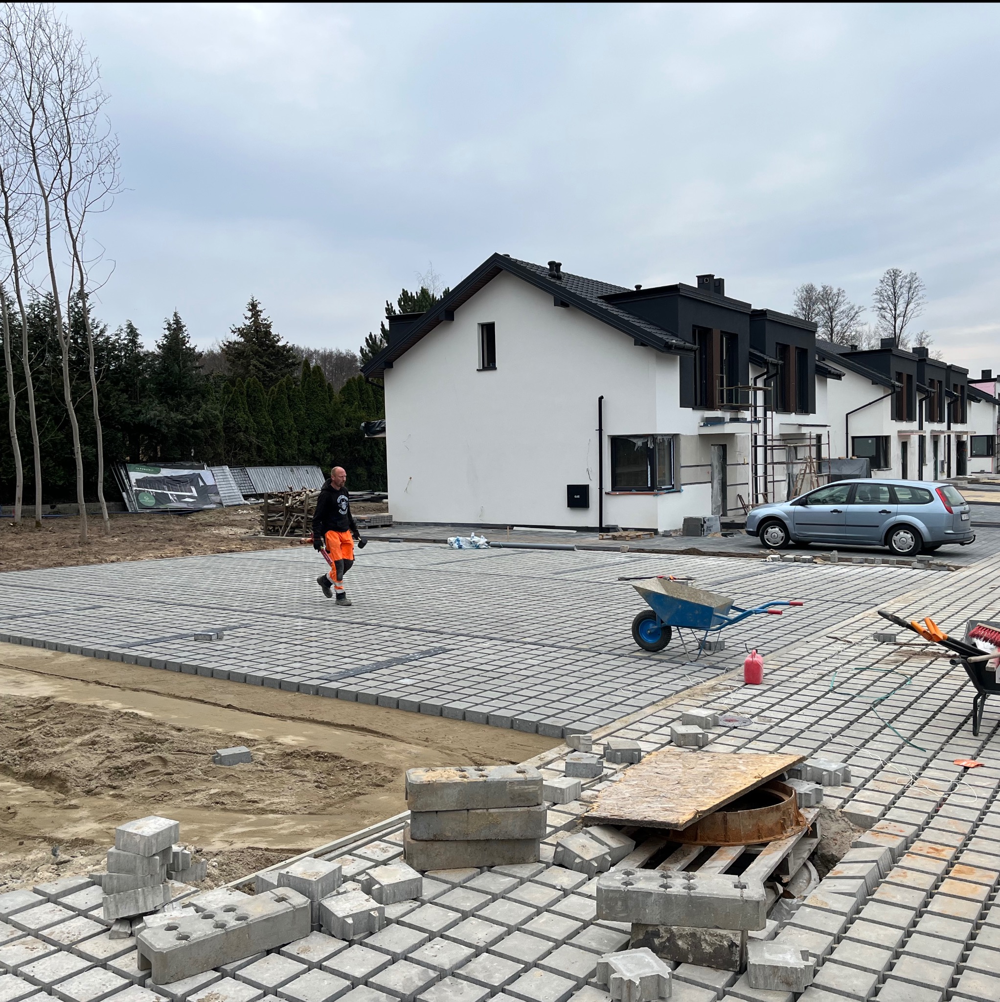 Układanie szarej kostki brukowej na posesji przed nowoczesnymi domami. Widoczny pracownik w odblaskowej odzieży, taczka i narzędzia. Częściowo ułożona nawierzchnia.