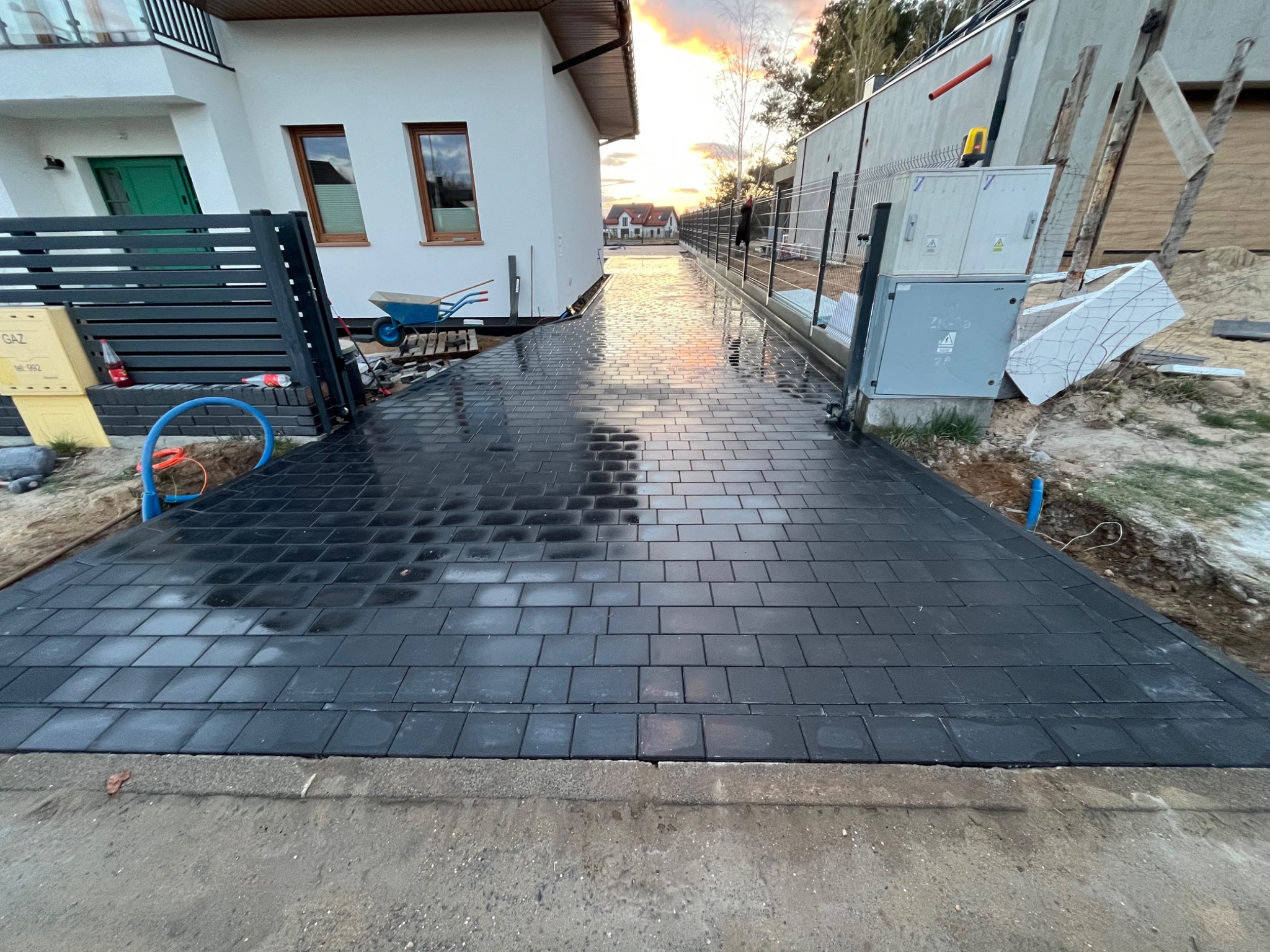 Świeżo ułożona, czarna kostka brukowa na podjeździe w Łodzi, odbijająca zachodzące słońce. Widoczne detale: skrzynka gazowa, brama wjazdowa i fragmenty domu.