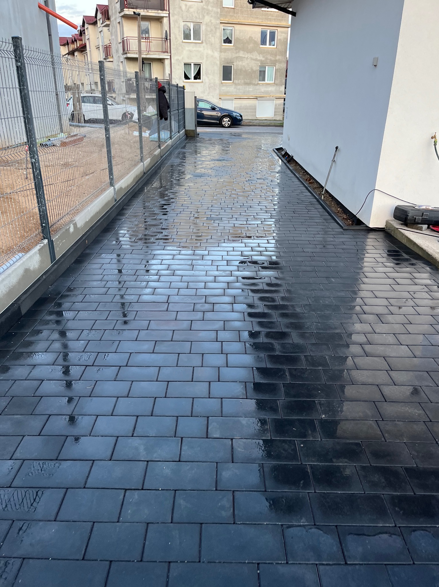 Świeżo ułożona, mokra, czarna kostka brukowa na podjeździe, odbijająca światło. Widoczny fragment ogrodzenia i budynków w tle. Minimalistyczny design.