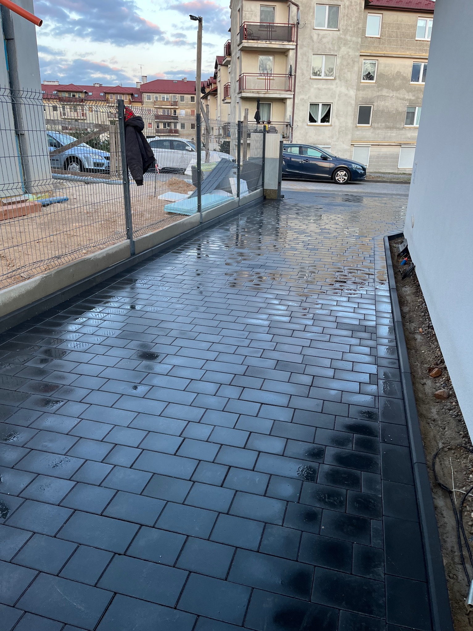 Świeżo ułożona, mokra, czarna kostka brukowa wzdłuż ogrodzenia posesji. Widoczne budynki w tle i elementy instalacji elektrycznej przy ścianie.