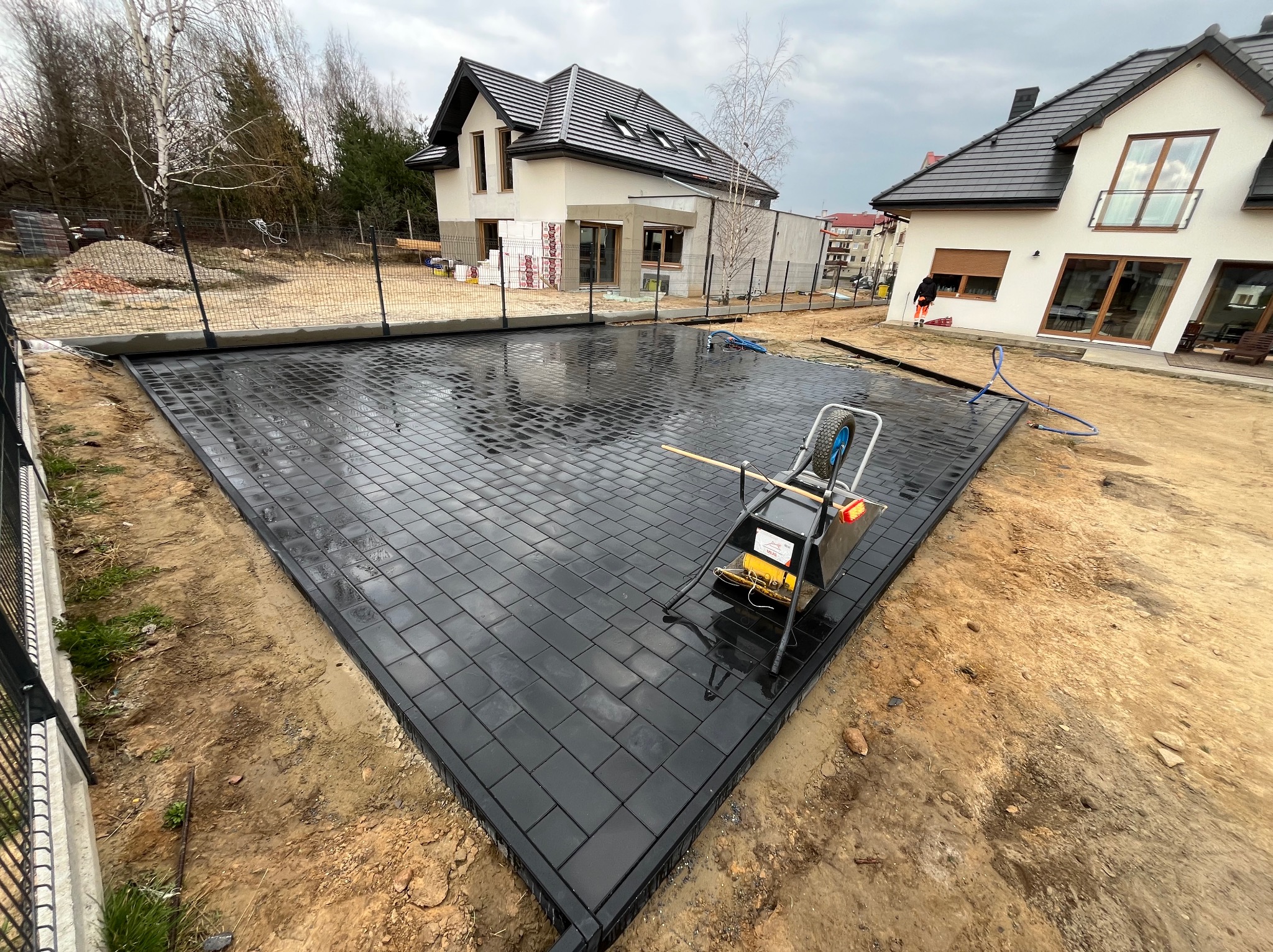 Ułożona, czarna kostka brukowa na posesji, widoczna ubijarka. Świeżo wykonana nawierzchnia, mokra od deszczu, otoczona ziemią i ogrodzeniem. W tle domy jednorodzinne.