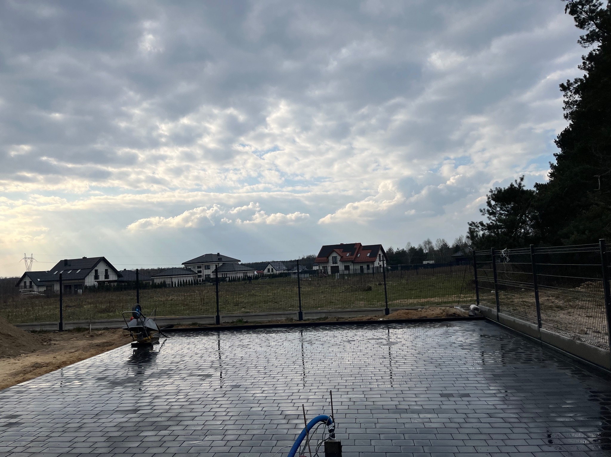 Świeżo ułożona, ciemnoszara kostka brukowa na posesji z widokiem na domy w tle. Częściowo widoczna zagęszczarka i niebieski wąż. Pochmurne niebo.