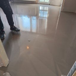 Wojciech Piatek - Samopoziomująca wylewka podłogowa w jasnym odcieniu szarości, odbijająca światło okna. Widoczne fragmenty ubrania i obuwie osoby wykonującej prace.