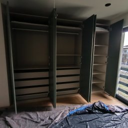 Wojciech Piatek - Wnętrze szafy wnękowej z otwartymi drzwiami, widoczne półki i szuflady. Podłoga zabezpieczona folią malarską. Widok z dołu na zamontowaną szafę.