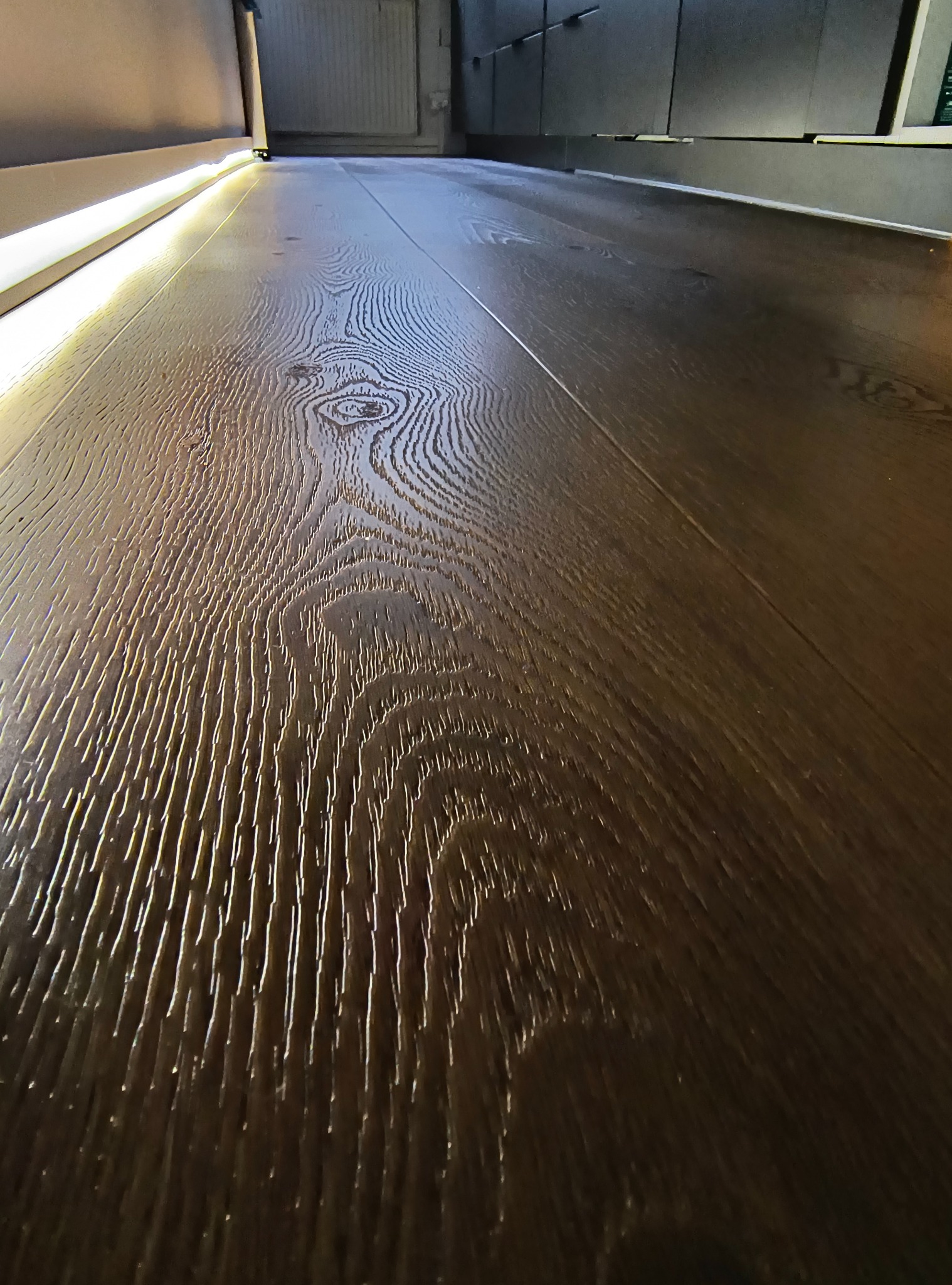 Położony parkiet z ciemnego drewna z widoczną fakturą i subtelnym oświetleniem ledowym przy ścianie. Perspektywa z dołu podkreśla głębię i wzór drewna.