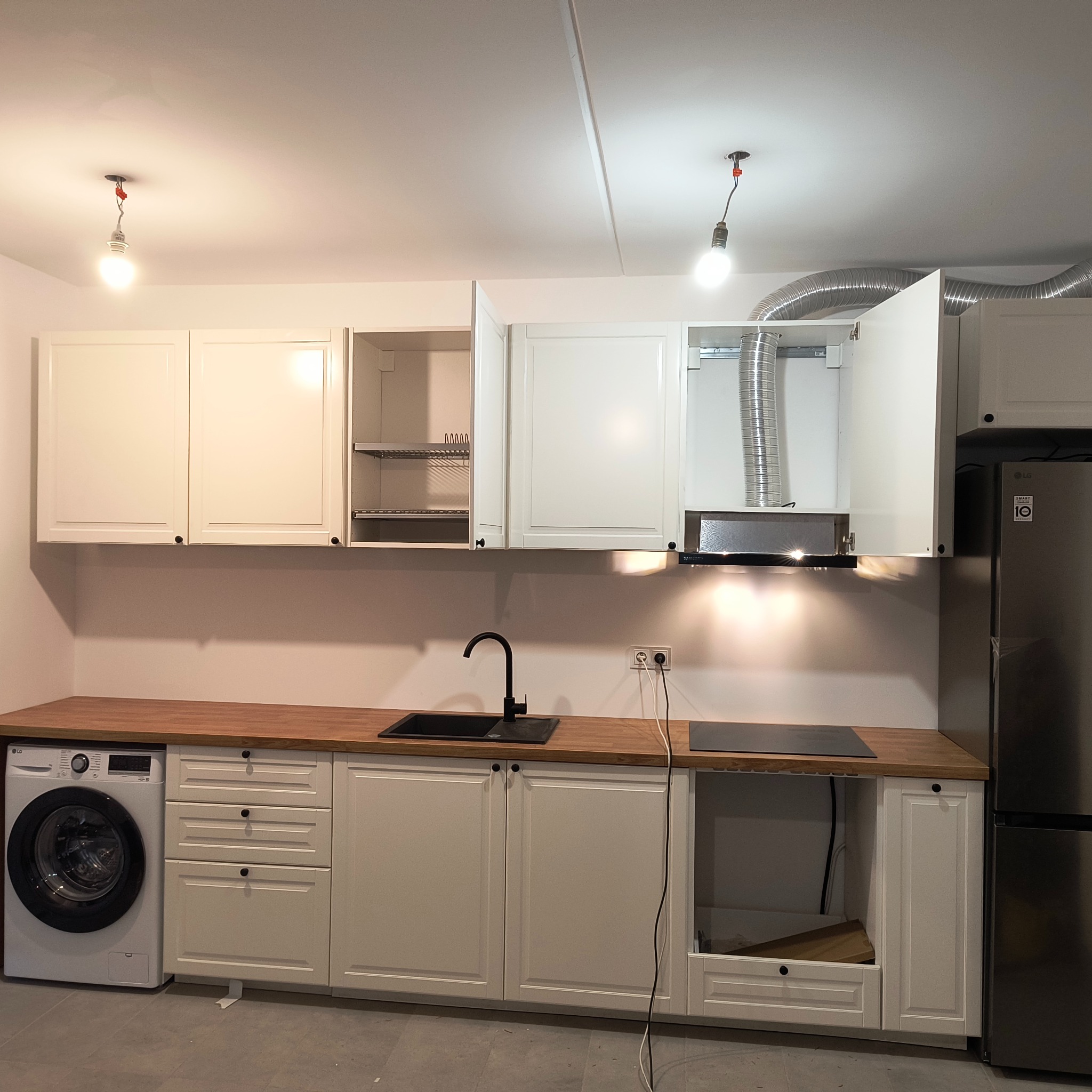 Jasna kuchnia z drewnianym blatem, czarnym zlewem i sprzętem AGD. Widoczne niedokończone szafki i wiszące żarówki, sugerujące etap montażu.