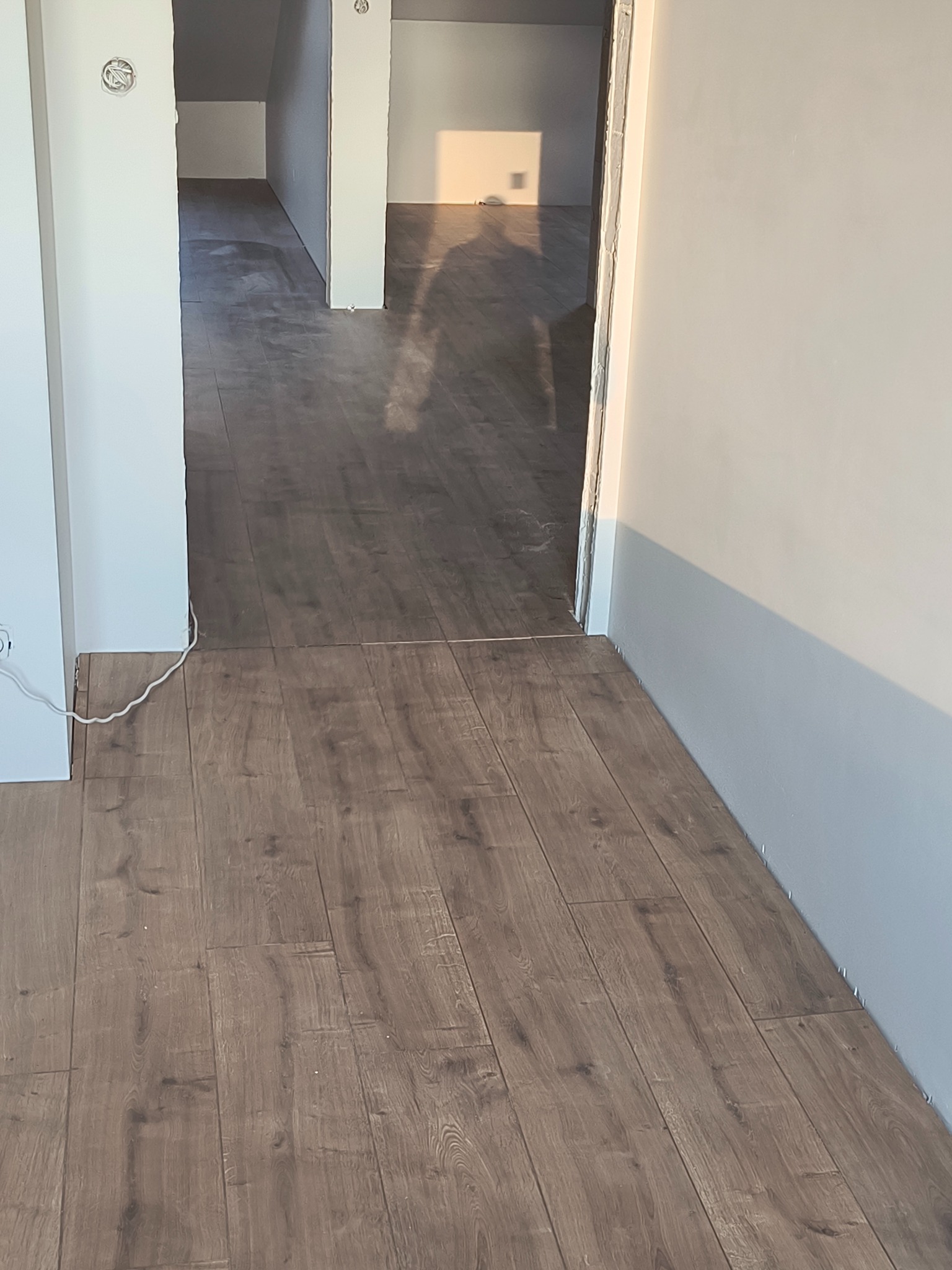 Ułożone panele podłogowe w korytarzu, widoczne przejścia do innych pomieszczeń. Ujęcie z niskiej perspektywy, podkreślające strukturę paneli i jasne ściany.