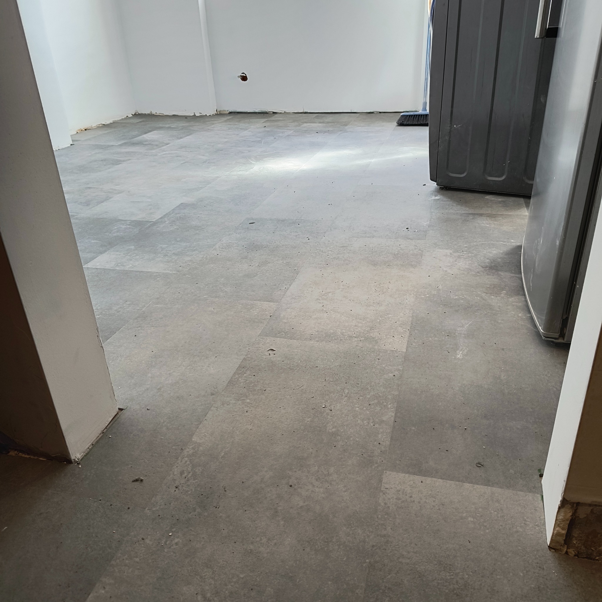 Szare panele podłogowe ułożone w pomieszczeniu, widoczne fragmenty ścian i pralka. Podłoga wykończona, gotowa do użytkowania, z widoczną strukturą paneli.