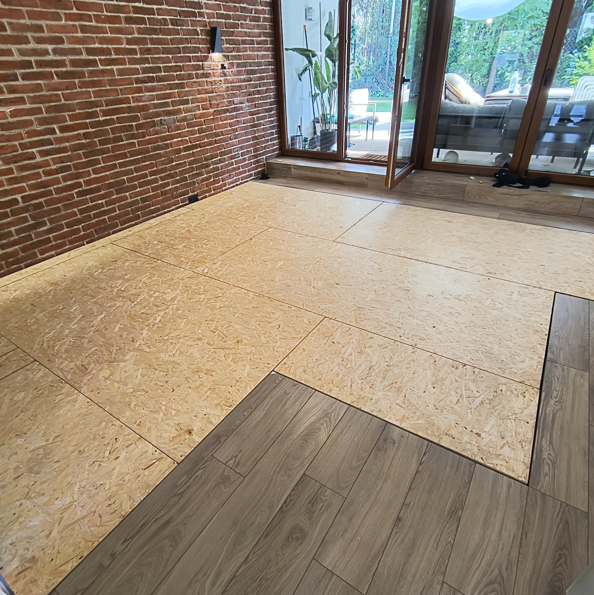 Podkład z płyt OSB pod podłogę, częściowo pokryty panelami imitującymi drewno, na tle ceglanej ściany i otwartych drzwi balkonowych.