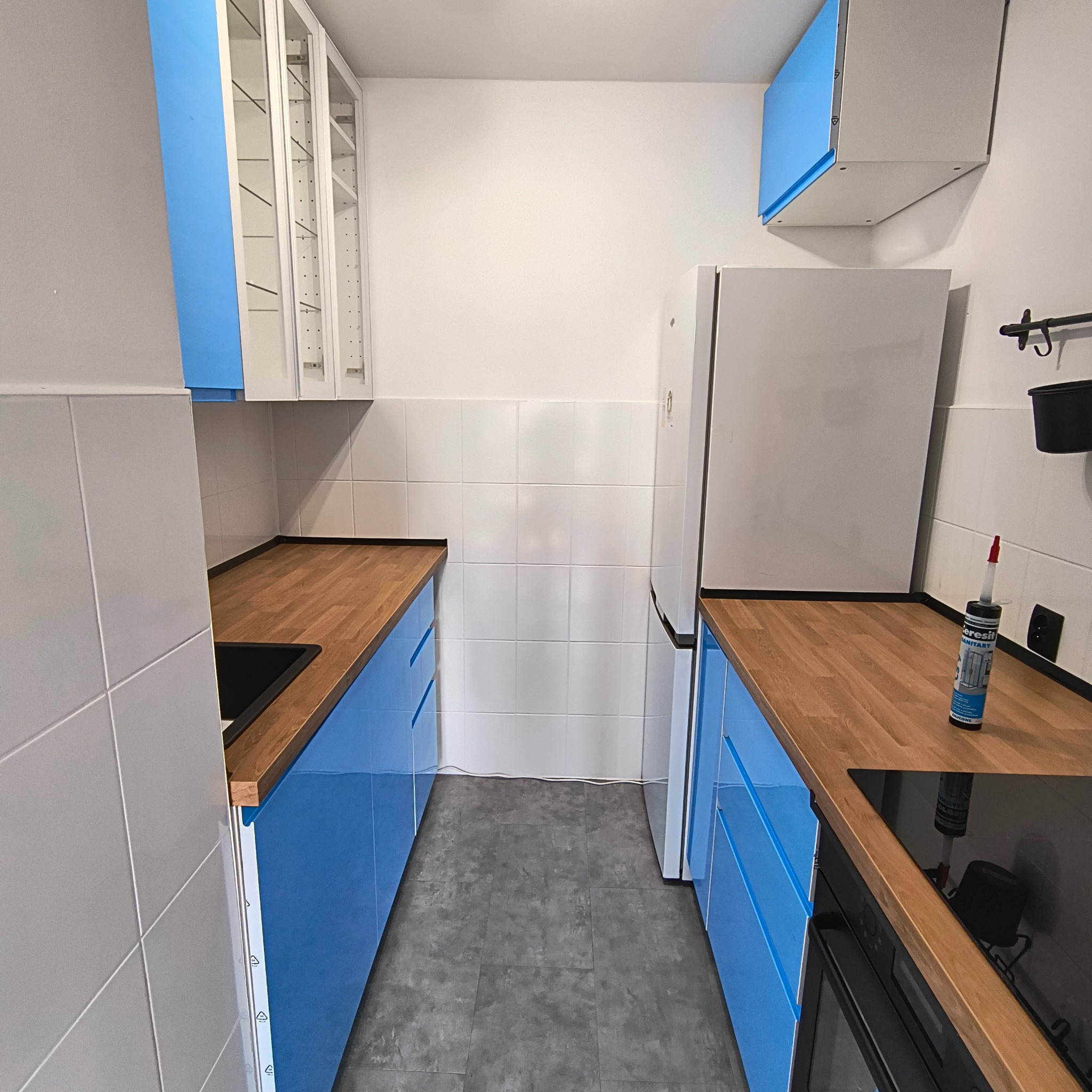 Wąska kuchnia z niebieskimi szafkami, drewnianym blatem i białymi płytkami. Tubka silikonu na blacie. Ujęcie z poziomu podłogi, podkreślające głębię.