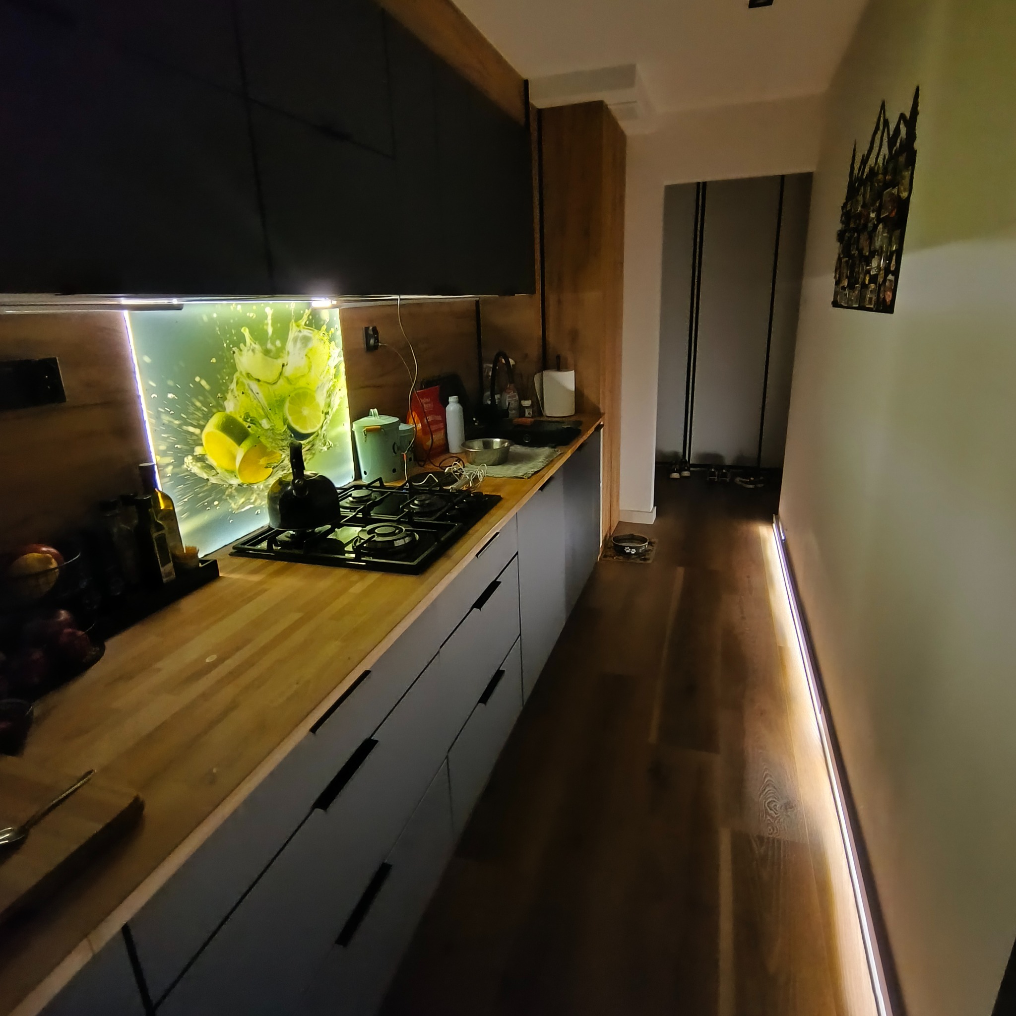 Nowoczesna kuchnia z szarymi szafkami, drewnianym blatem i podświetlanym panelem z motywem owoców. Parkiet z oświetleniem LED wzdłuż ściany.