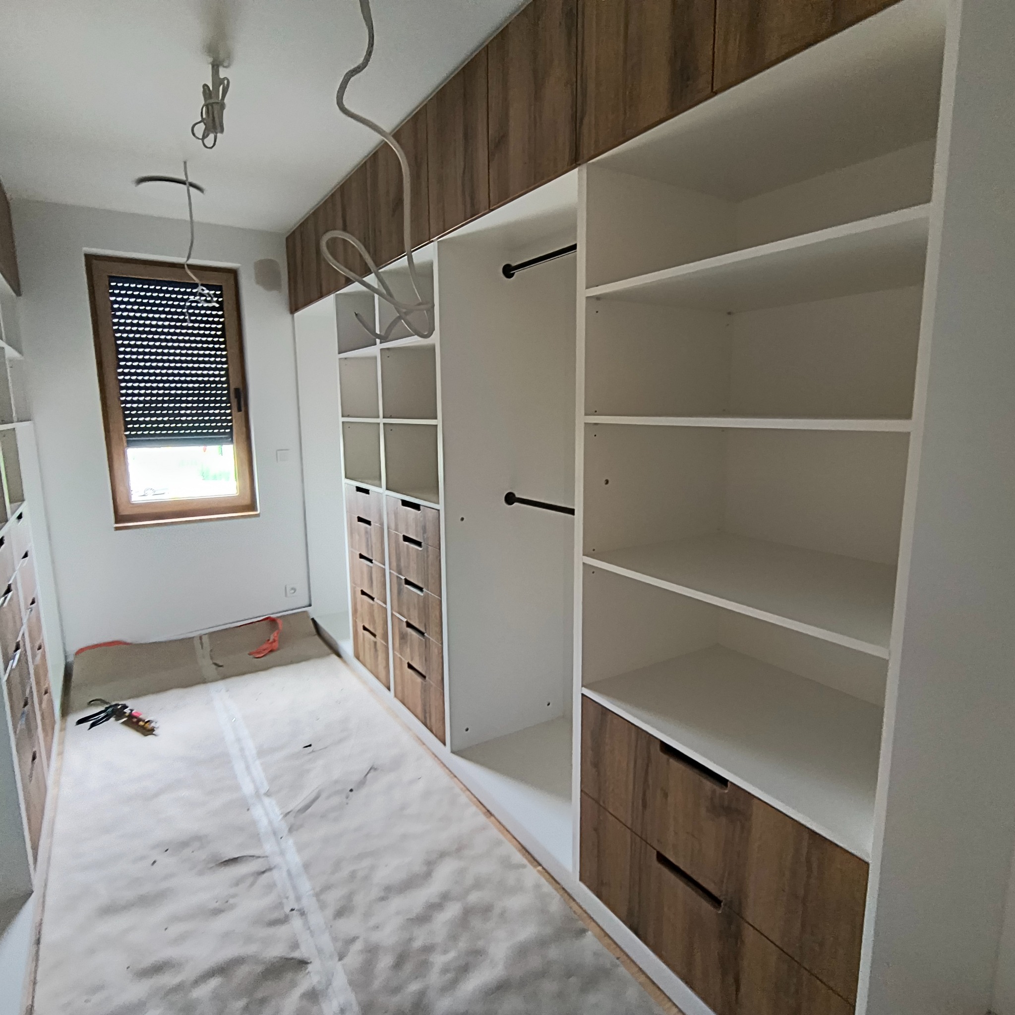 Realizacja zabudowy garderoby z drewnianymi akcentami w Tychach. Widoczne półki, szuflady i drążki na ubrania. Podłoga zabezpieczona folią malarską.