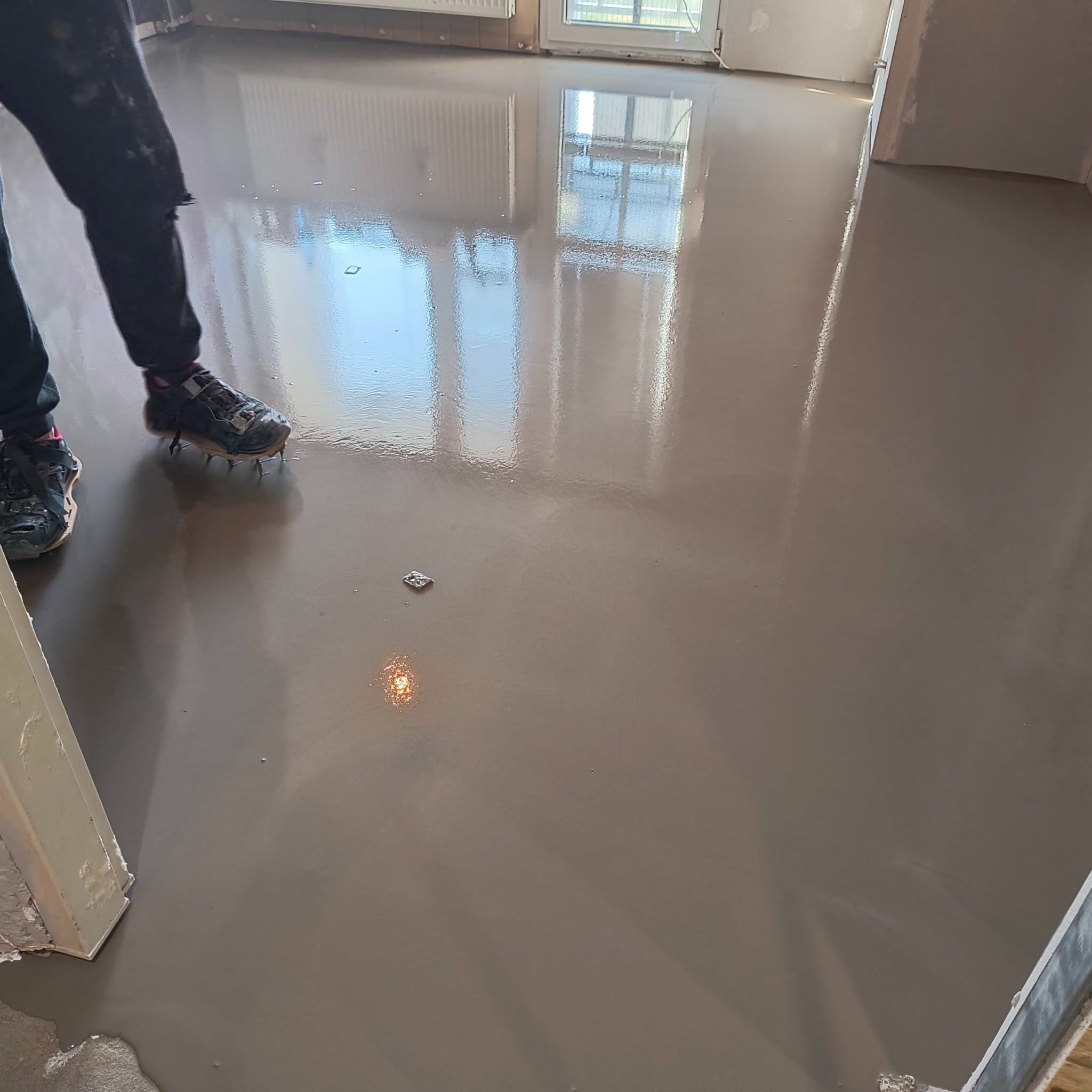 Samopoziomująca wylewka podłogowa w jasnym odcieniu szarości, odbijająca światło okna. Widoczne fragmenty ubrania i obuwie osoby wykonującej prace.