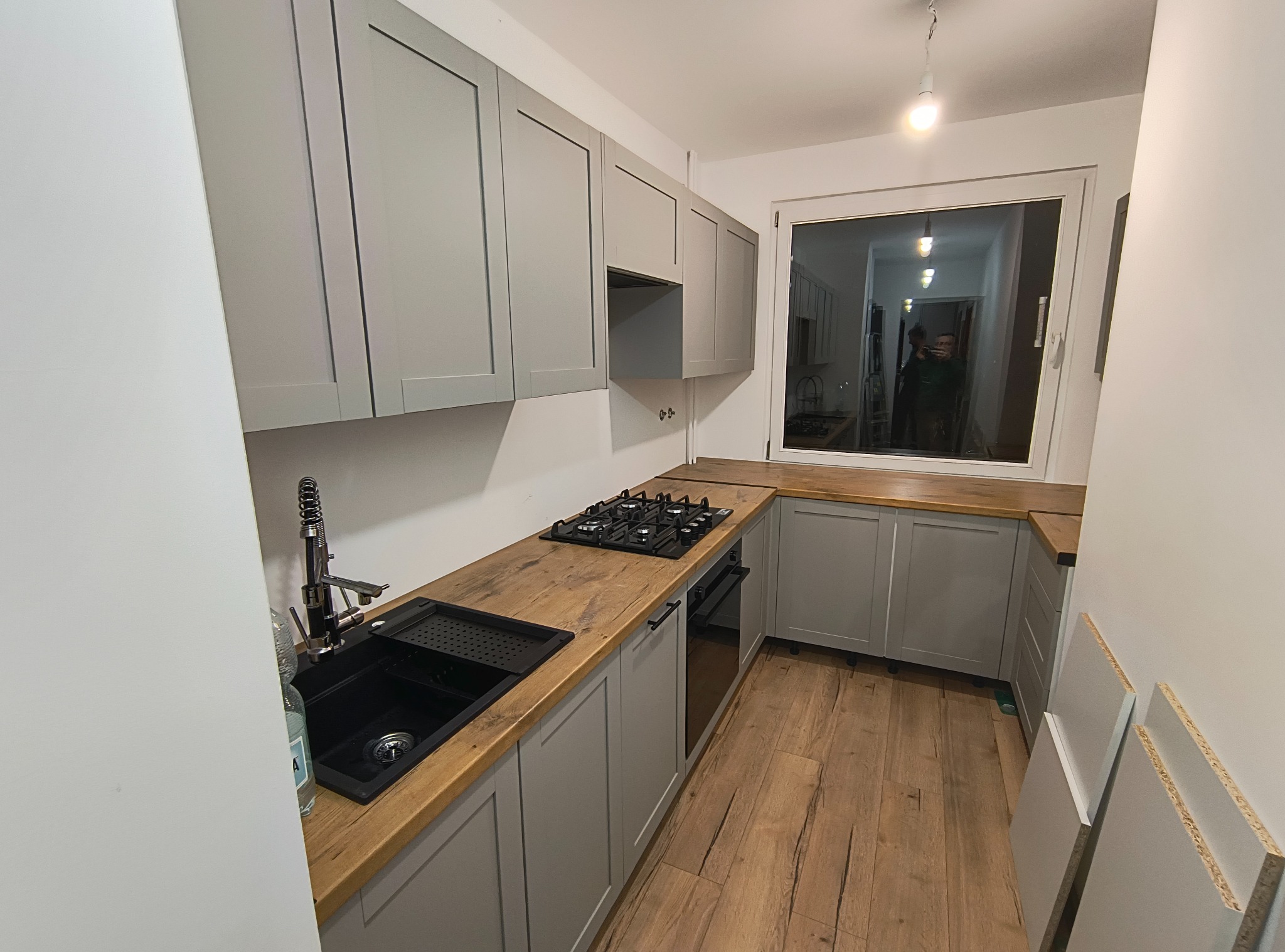 Jasnoszara zabudowa kuchenna z drewnianym blatem, czarnym zlewem i płytą gazową. Widok na okno z odbiciem wnętrza. Podłoga z paneli imitujących drewno.