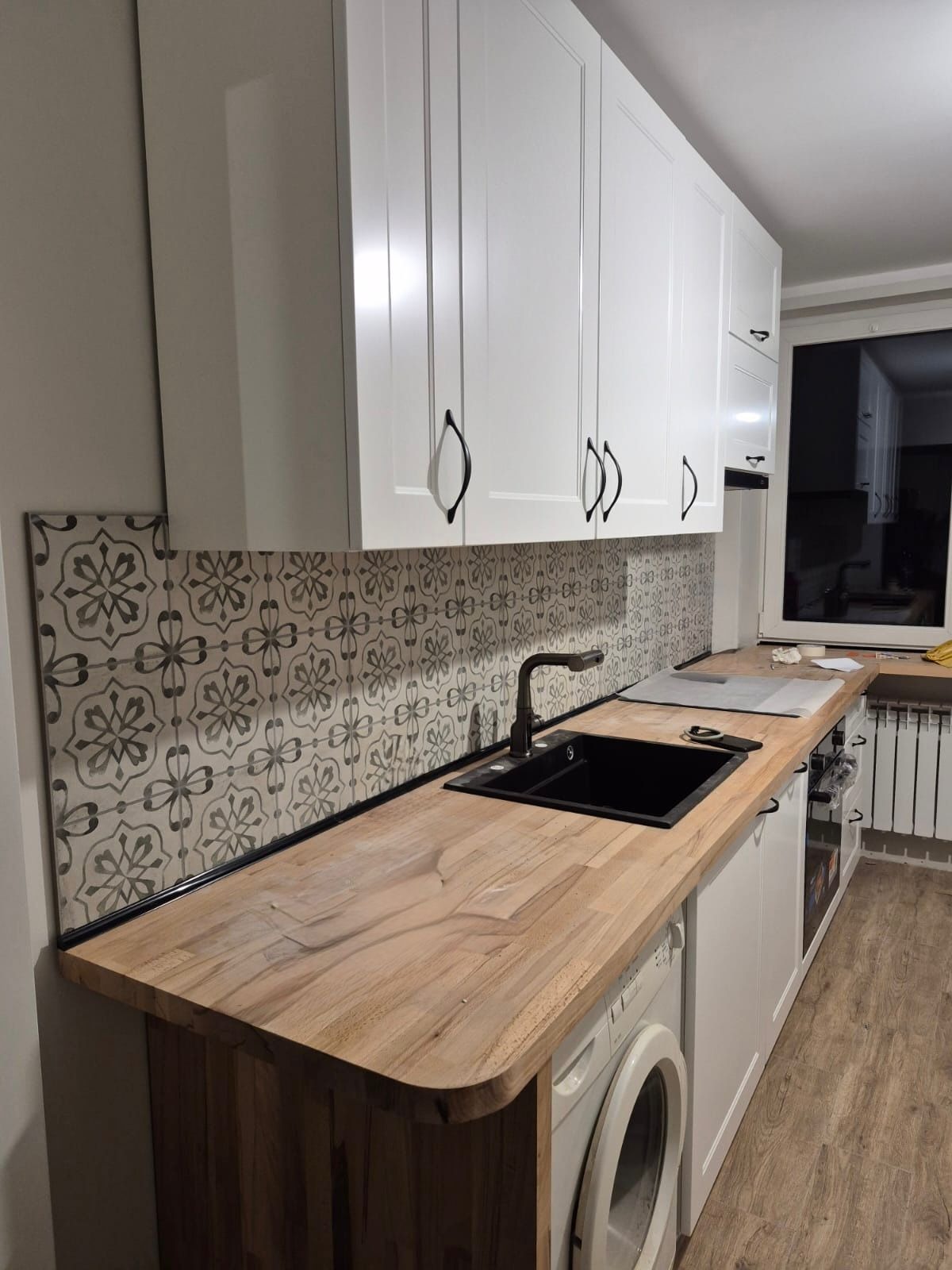 Białe szafki kuchenne z czarnymi uchwytami i drewnianym blatem. Nad blatem wzorzyste płytki. W tle okno. Widoczna pralka pod blatem.