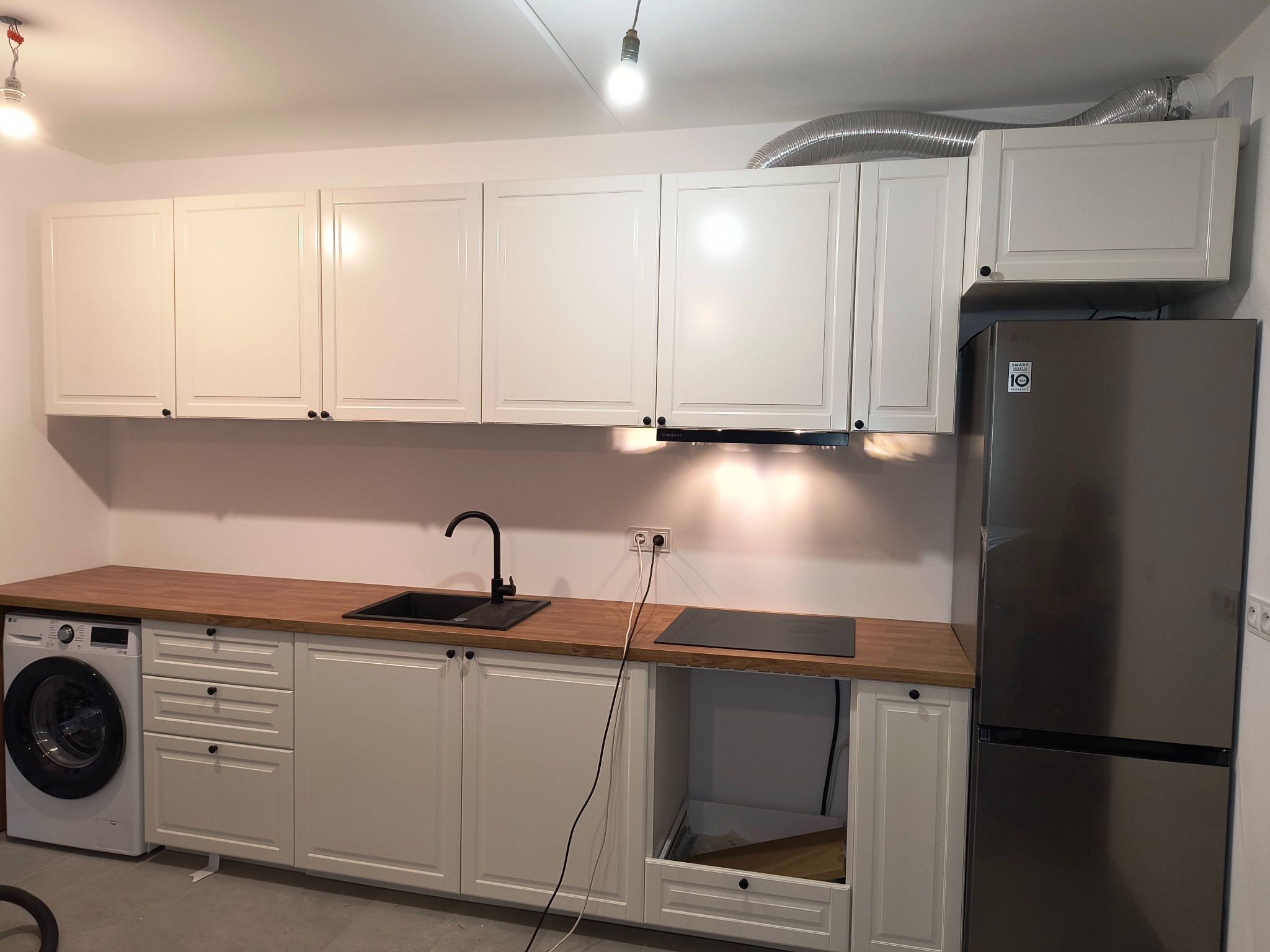 Biała zabudowa kuchenna z drewnianym blatem, czarnym zlewem i płytą. Obok pralka i lodówka. Widoczny fragment wentylacji. Oświetlenie punktowe.