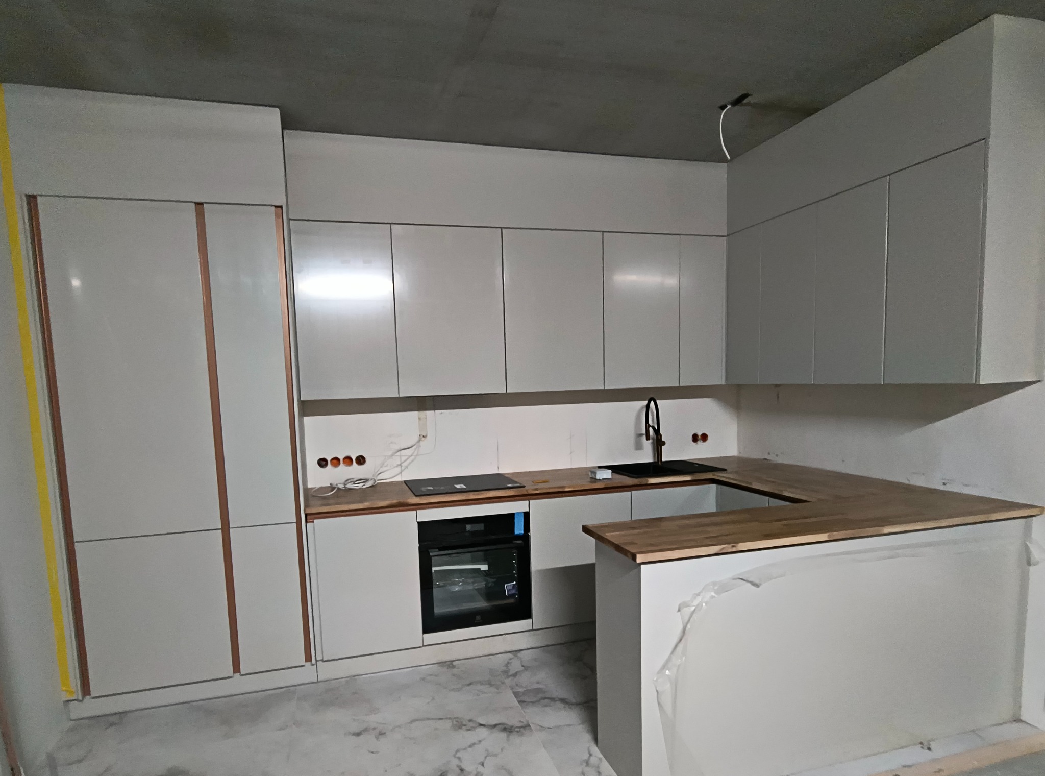 Nowoczesna, jasna kuchnia z białymi szafkami i drewnianym blatem, jeszcze w trakcie wykończenia. Widoczne gniazdka i marmurowa podłoga.