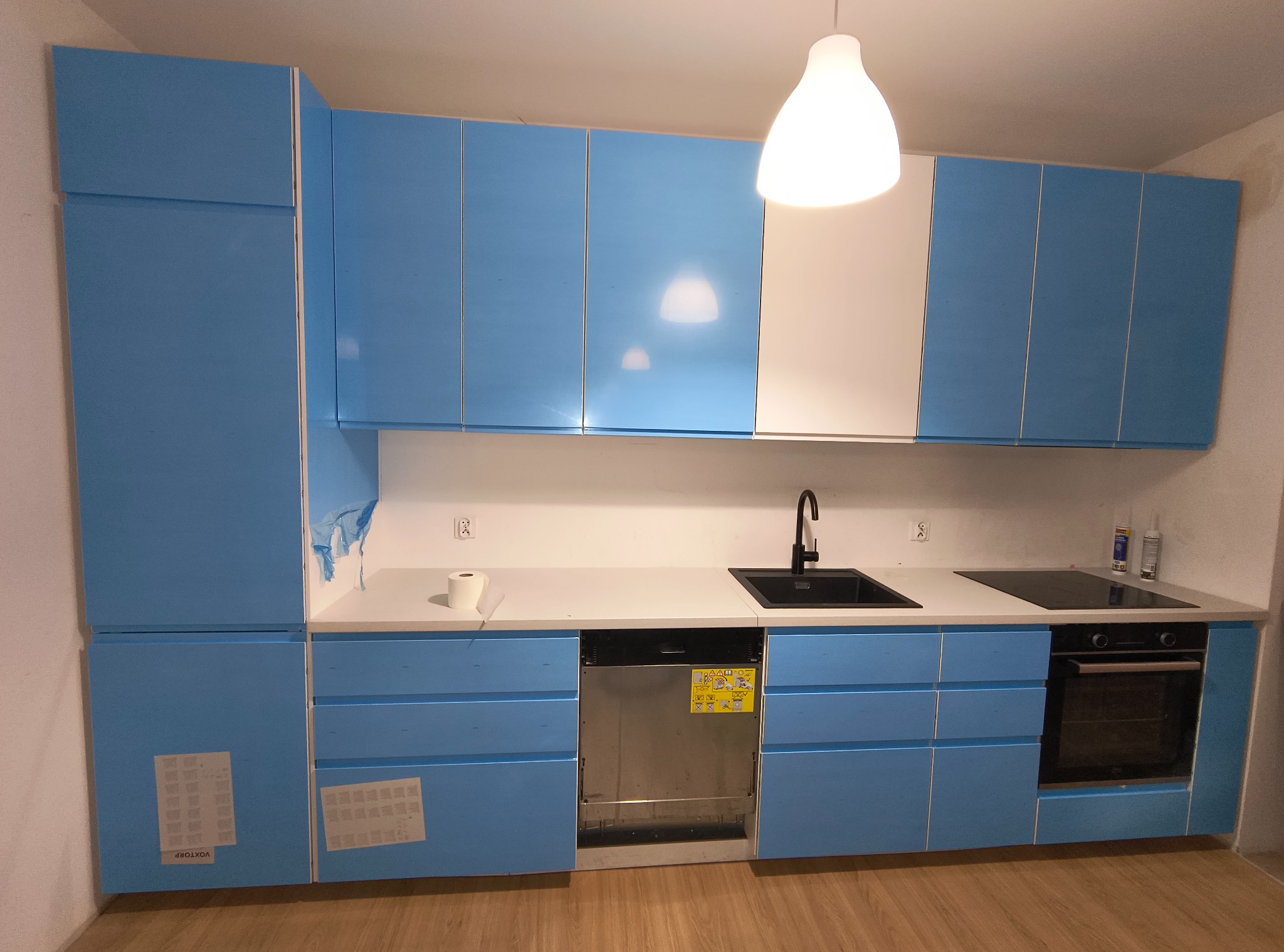 Niebieska zabudowa kuchenna z białym blatem i sprzętami AGD, częściowo jeszcze zabezpieczona folią ochronną. Widoczna lampa sufitowa i drewniana podłoga.