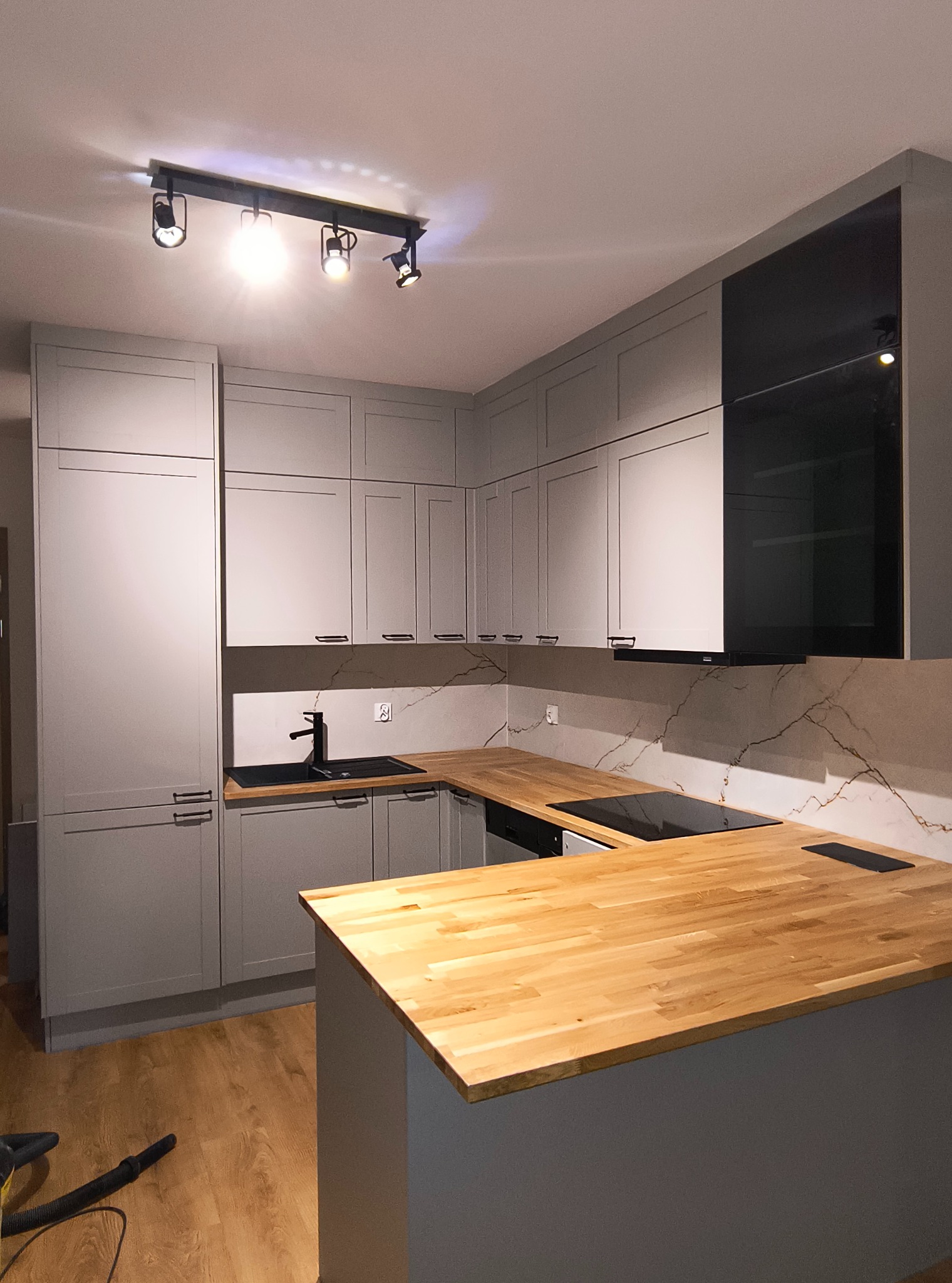 Szara kuchnia z drewnianym blatem i czarnymi akcentami, oświetlona reflektorami. Widoczny fragment podłogi i odkurzacza. Nowoczesny design.