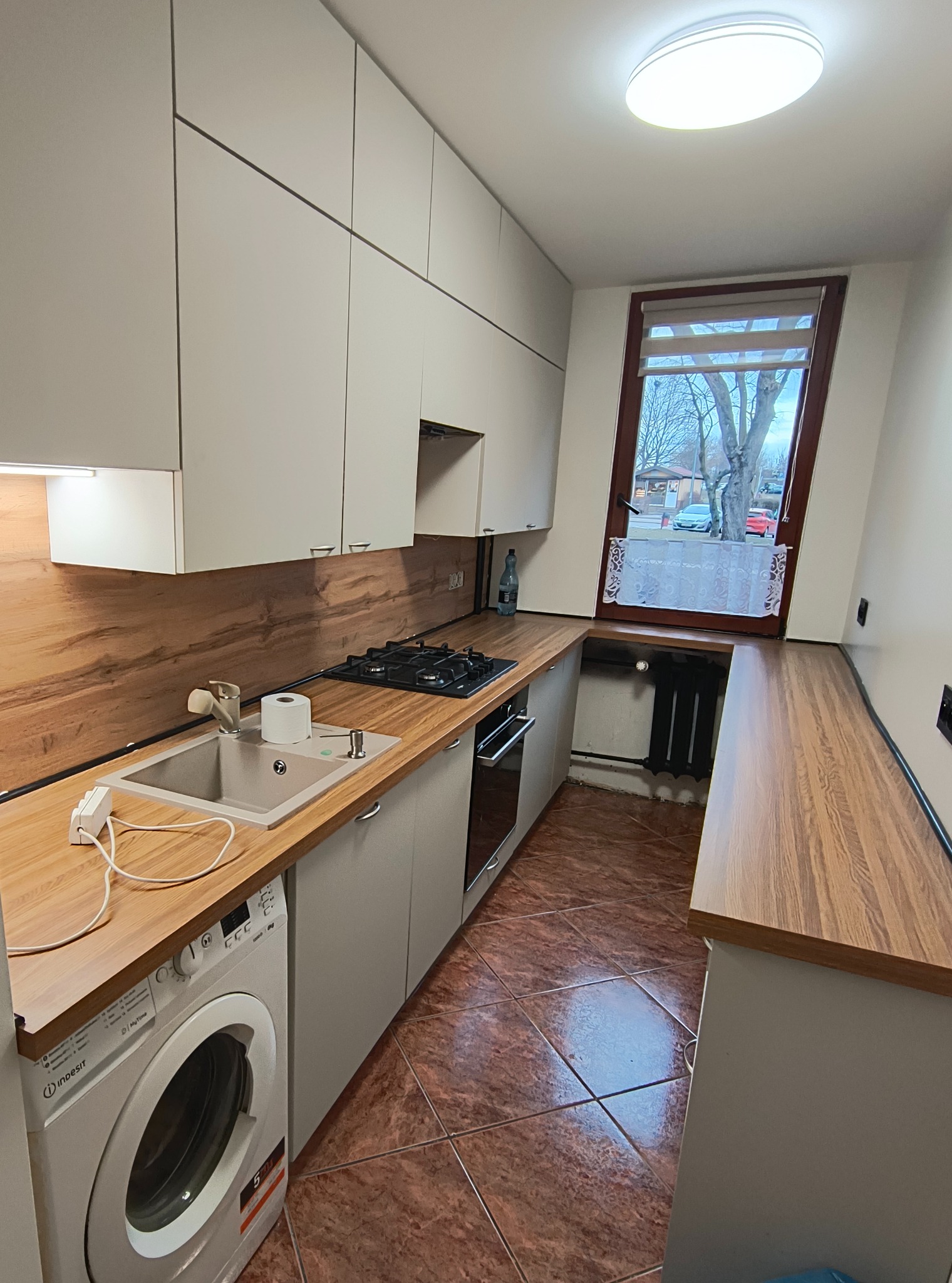 Nowoczesna kuchnia z szarymi szafkami i drewnianym blatem. Widoczna pralka, zlew, płyta gazowa i okno z widokiem na zewnątrz. Brązowa podłoga z płytek.