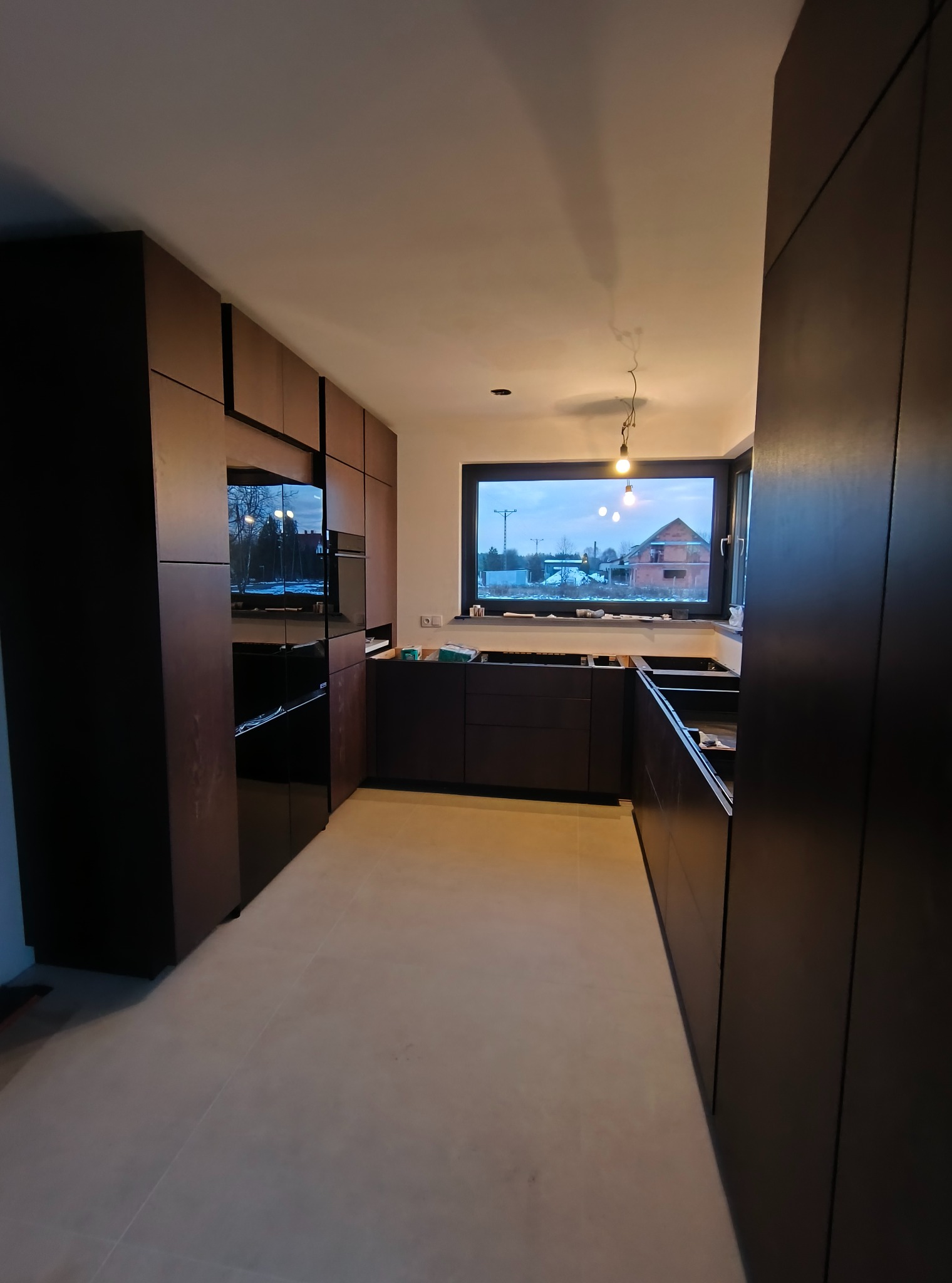 Ciemna, nowoczesna zabudowa kuchenna w trakcie montażu. Widoczne szafki bez blatów, okno z widokiem na zewnątrz. Minimalistyczny design, surowe wykończenie.