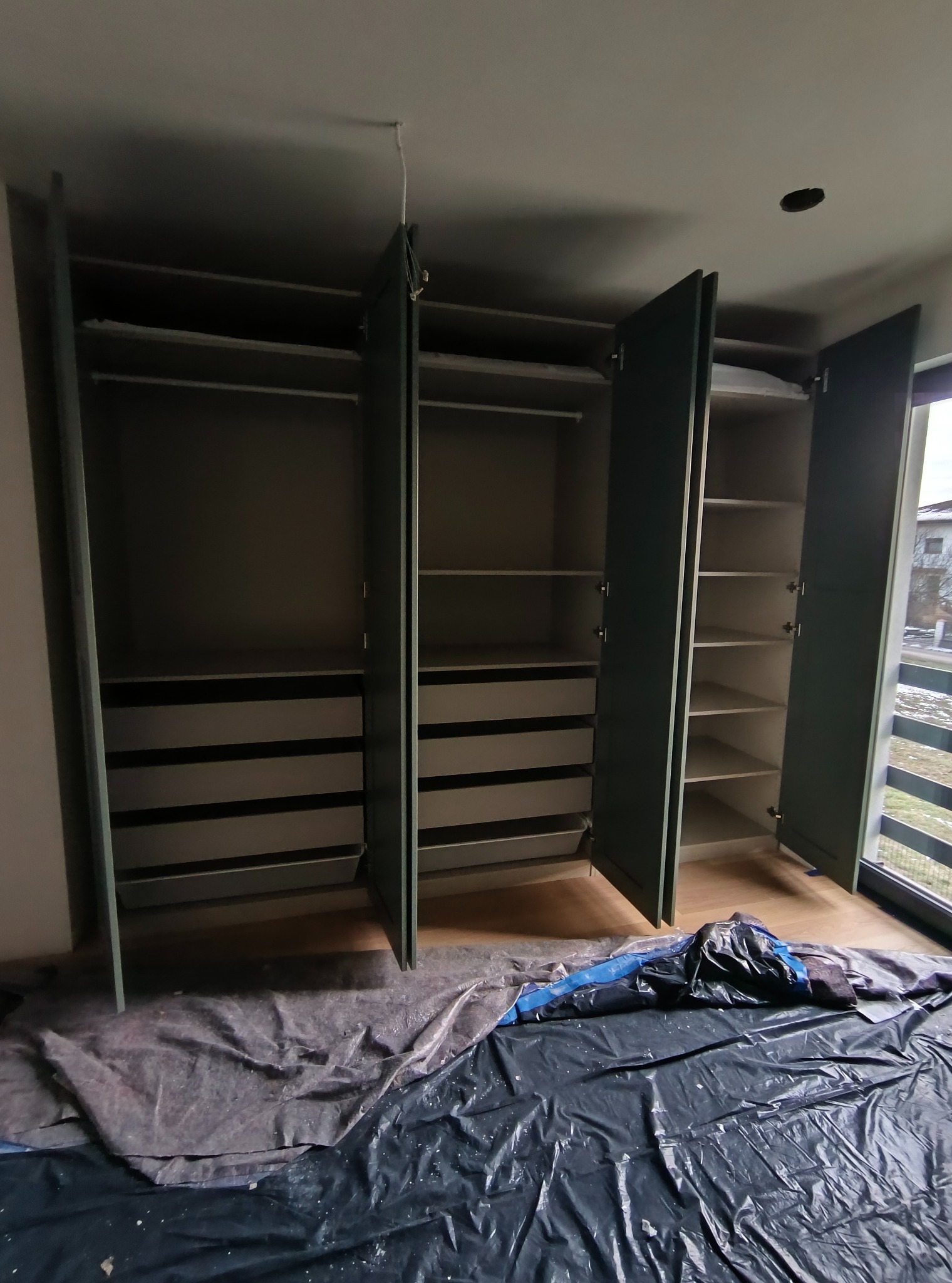 Wnętrze szafy wnękowej z otwartymi drzwiami, widoczne półki i szuflady. Podłoga zabezpieczona folią malarską. Widok z dołu na zamontowaną szafę.