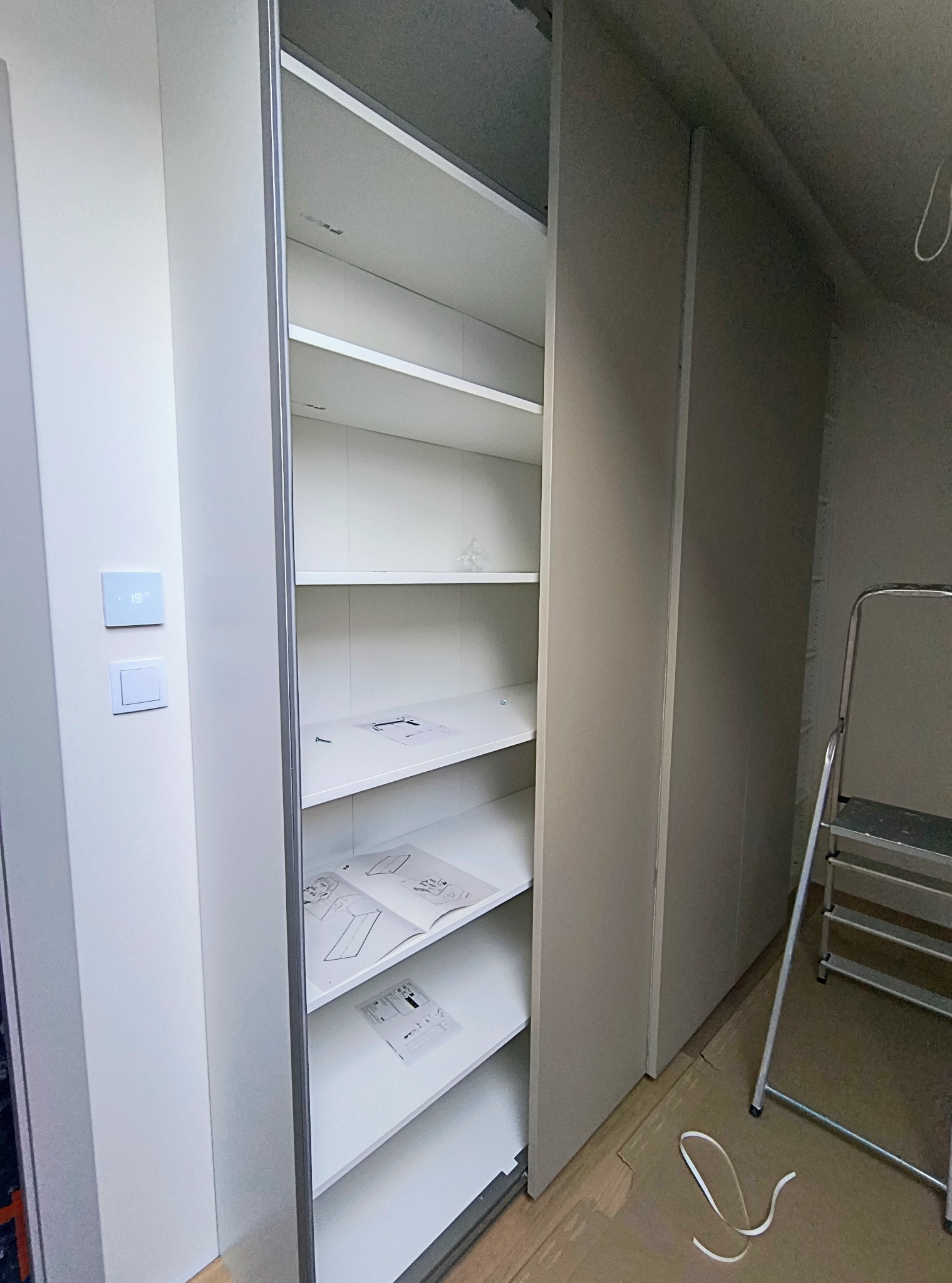 Biała szafa przesuwna z instrukcją montażu na półce, drabina oparta o ścianę. Widoczne elementy montażowe i niedoskonałości podłogi. Ujęcie z dołu, wnętrze w trakcie montażu.