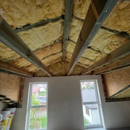 MIDEX - Ocieplone wełną mineralną poddasze z widocznymi belkami stropowymi i dwoma oknami. Widok na zewnątrz przez okna. Częściowo wykończone ściany.
