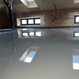 MIDEX - Wylany, mokry podkład podłogowy na poddaszu z oknami dachowymi. Widoczne kable i drewniana konstrukcja dachu. Podłoga odbija światło okien.