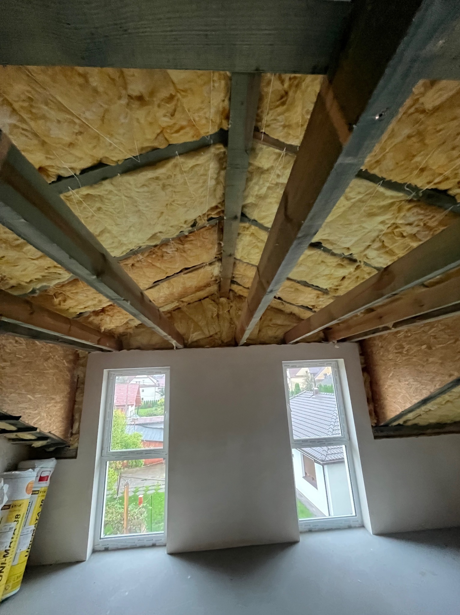 Ocieplone wełną mineralną poddasze z widocznymi belkami stropowymi i dwoma oknami. Widok na zewnątrz przez okna. Częściowo wykończone ściany.