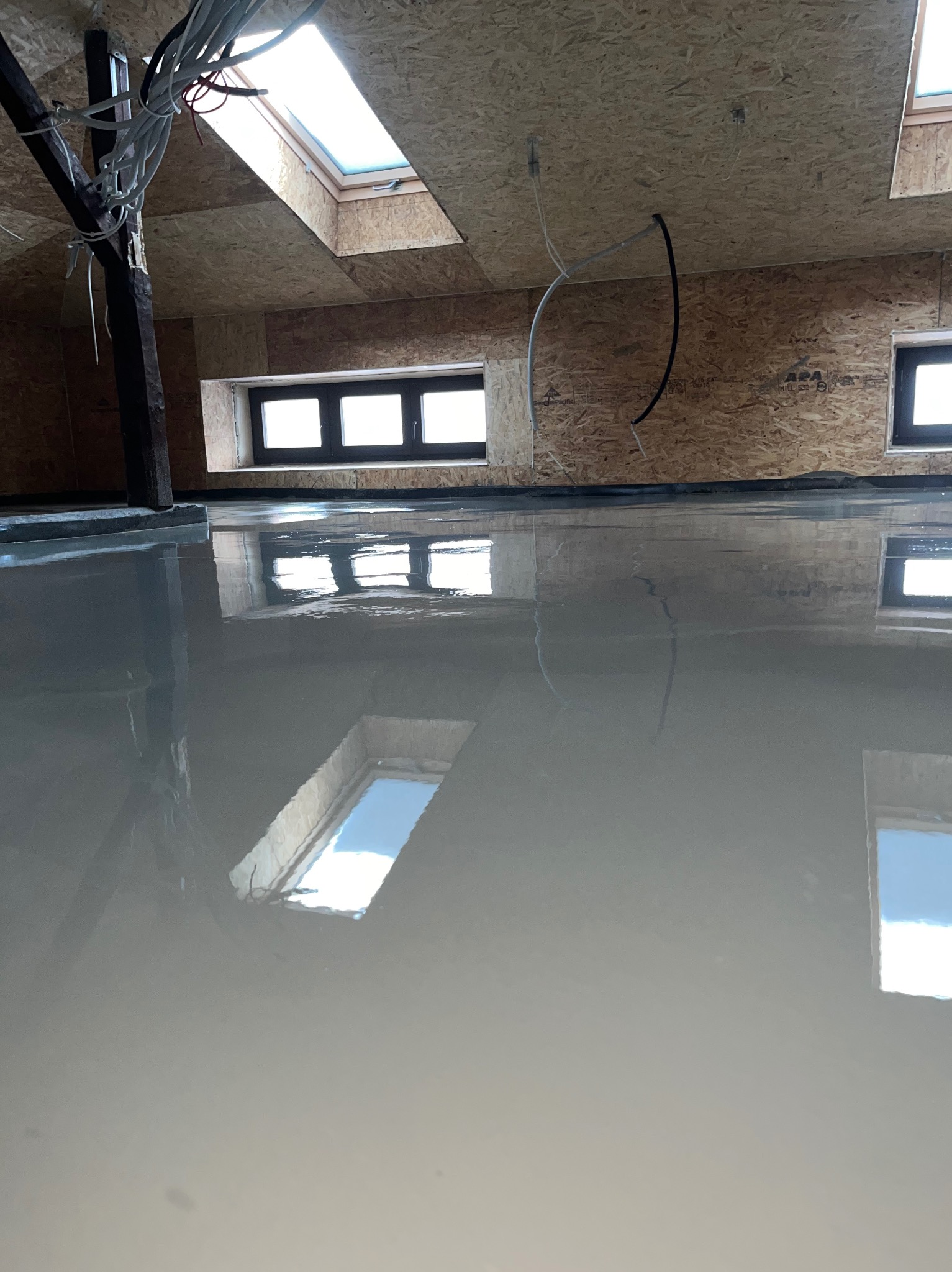 Wylany, mokry podkład podłogowy na poddaszu z oknami dachowymi. Widoczne kable i drewniana konstrukcja dachu. Podłoga odbija światło okien.