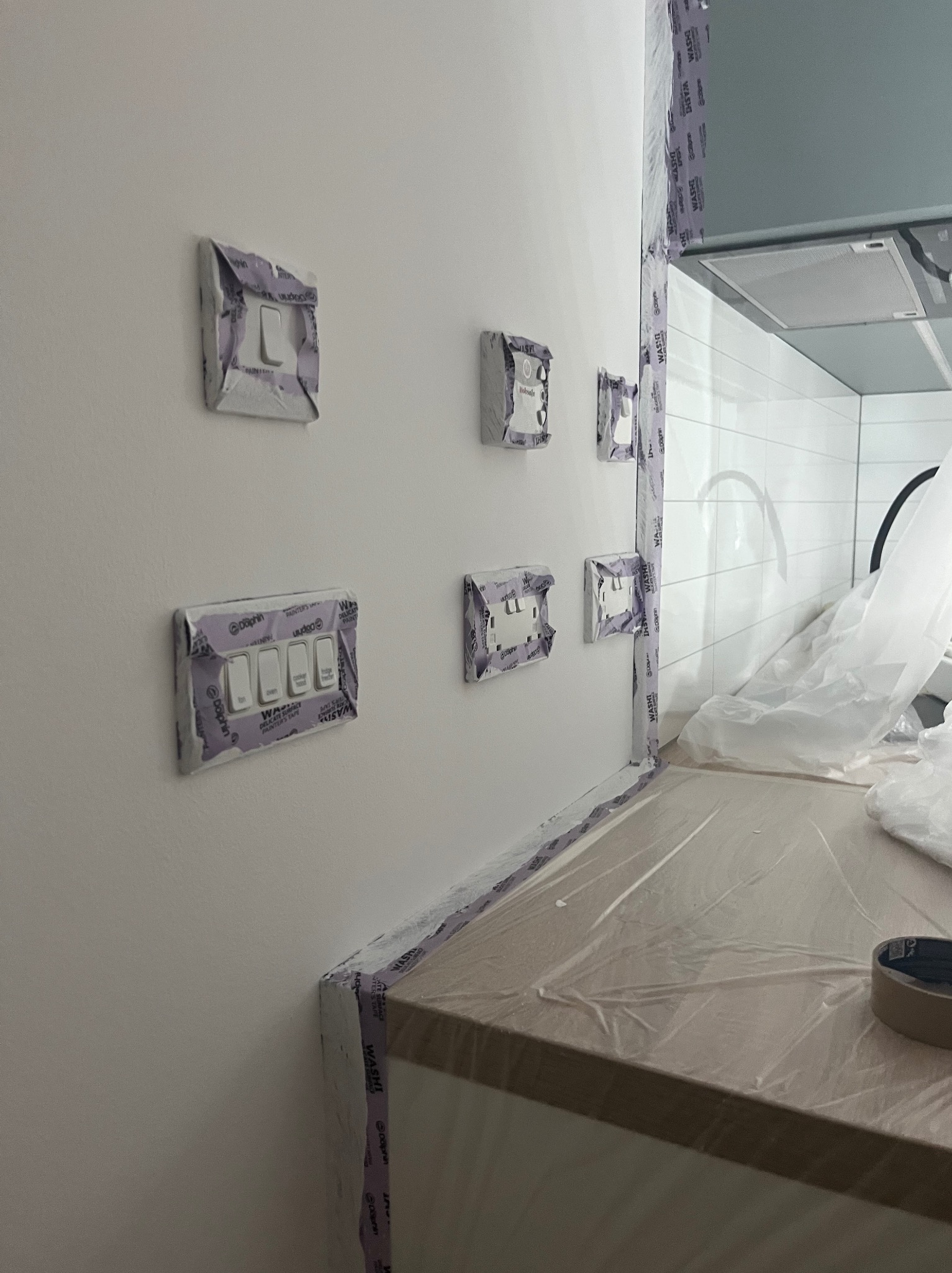 Przygotowanie do malowania: zabezpieczone włączniki i krawędzie taśmą malarską w pomieszczeniu z białymi płytkami. Ochrona przed zabrudzeniem farbą.