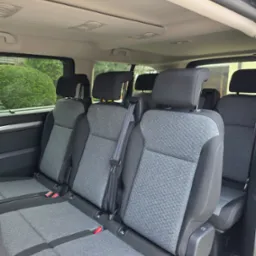 Wnętrze busa z trzema rzędami szarych siedzeń, widok z tyłu pojazdu. Widoczne zagłówki, pasy bezpieczeństwa i uchwyty ISOFIX.  Za oknami zieleń.