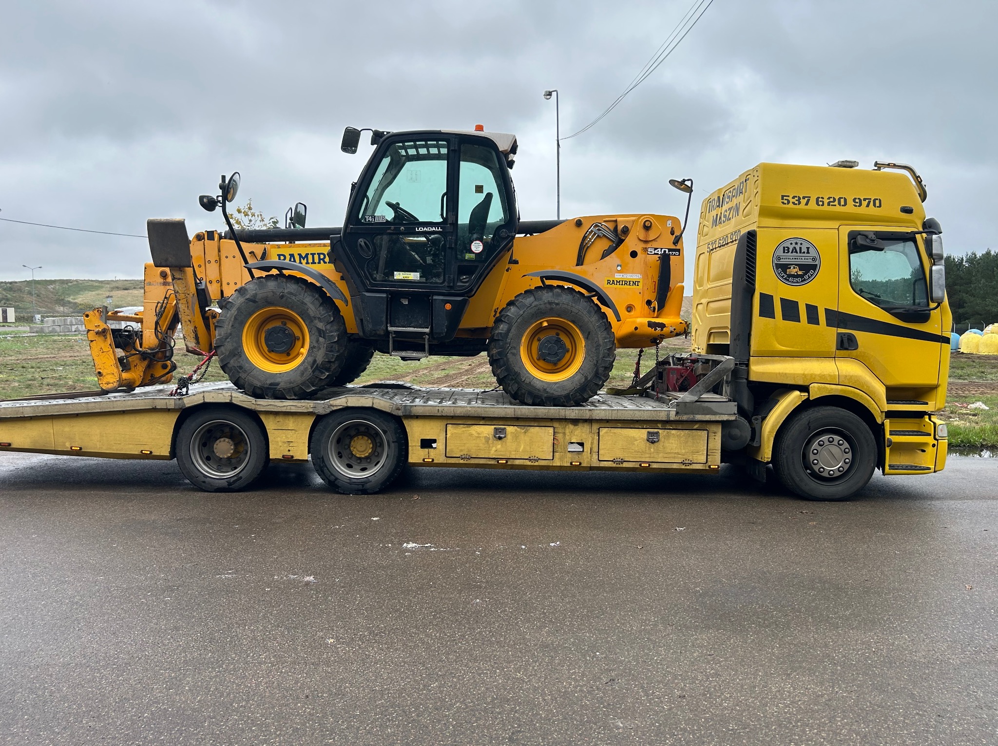 Żółta laweta z koparko-ładowarką Ramirent na platformie, widok z boku na szarej nawierzchni, pochmurne niebo w tle. Transport maszyny budowlanej.