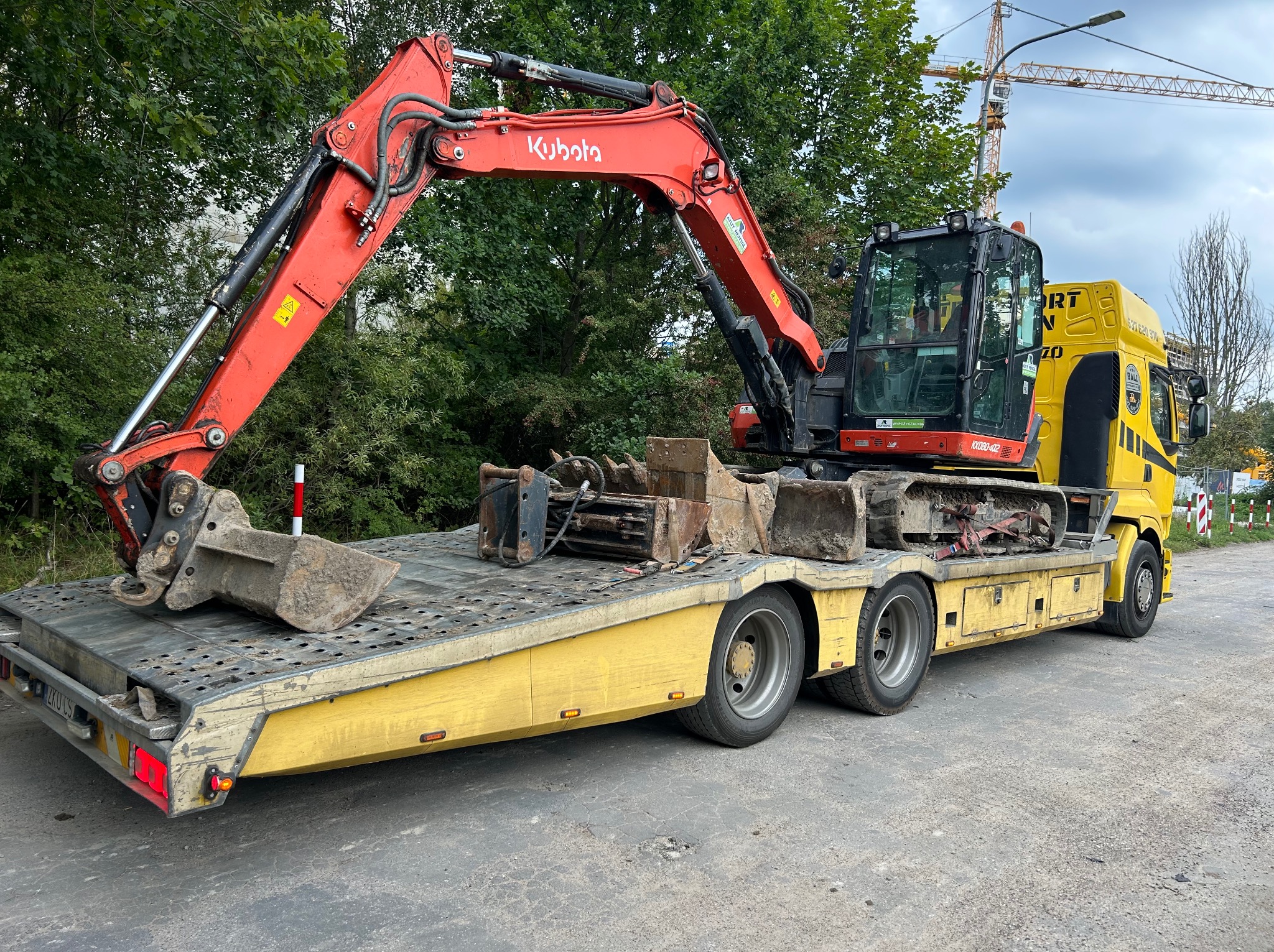 Żółta laweta z czerwoną mini koparką Kubota i łyżkami na platformie, na tle zieleni i żurawia budowlanego. Transport maszyn budowlanych.