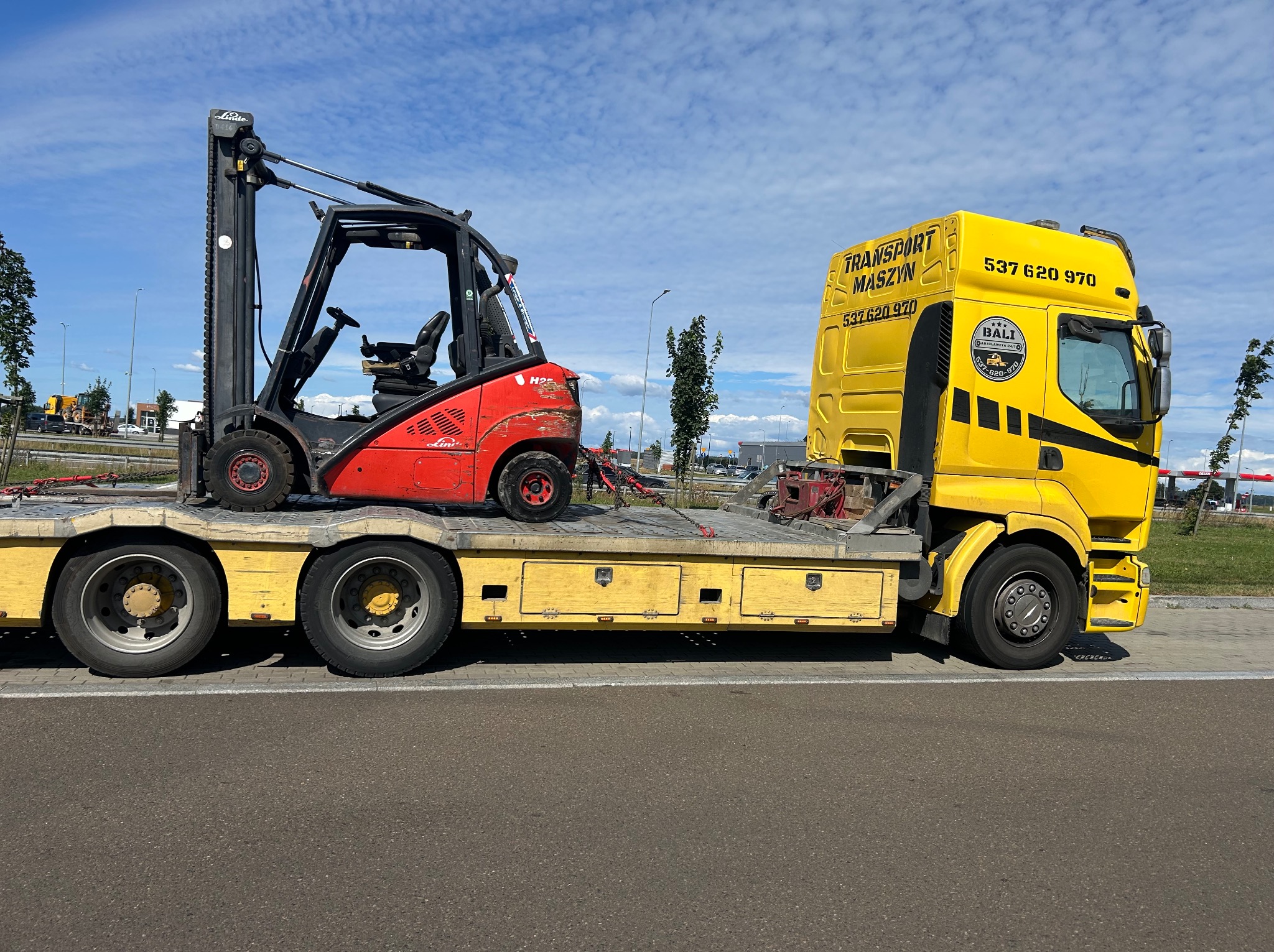 Żółta laweta z czerwonym wózkiem widłowym Linde na platformie, transport maszyn budowlanych. Widok z boku na asfaltowej drodze w słoneczny dzień.