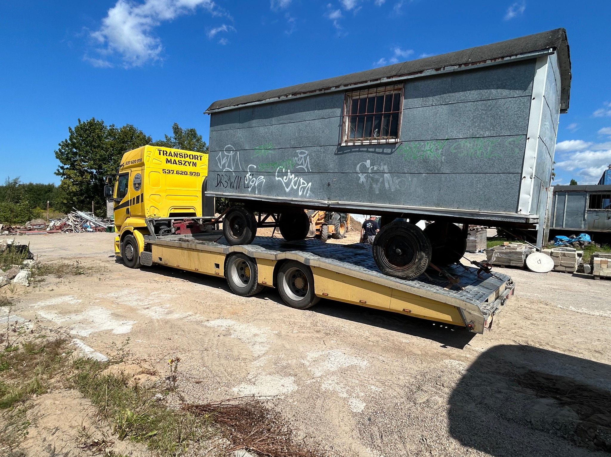 Żółta laweta transportuje szarą, blaszaną budkę z graffiti na platformie. Słoneczny dzień, błękitne niebo z chmurami w tle.