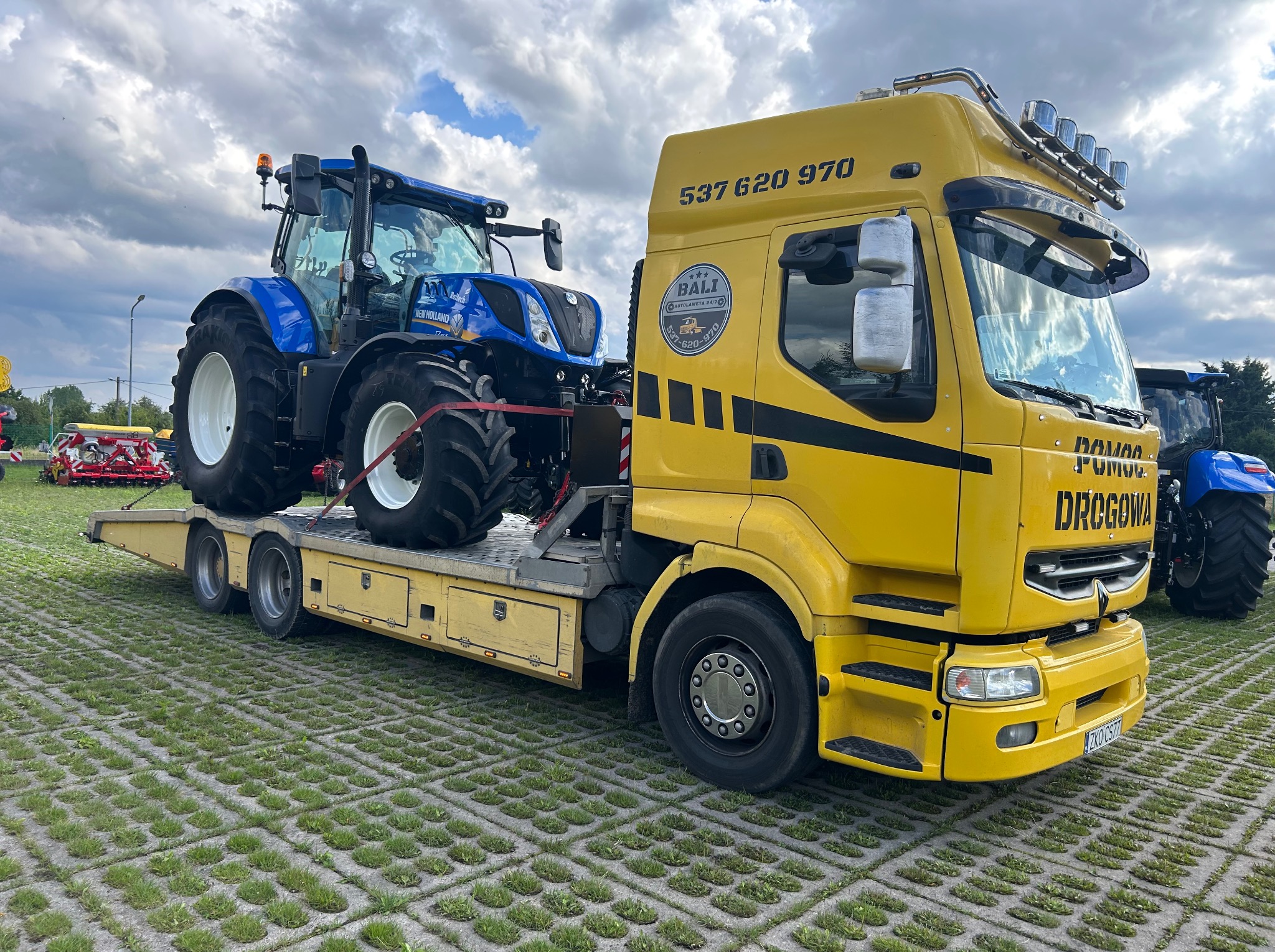 Żółta laweta z niebieskim traktorem na platformie w Starych Bielicach. Widoczne logo firmy 'BALI Autolaweta 24/7' i numer telefonu. Traktor zabezpieczony pasami.