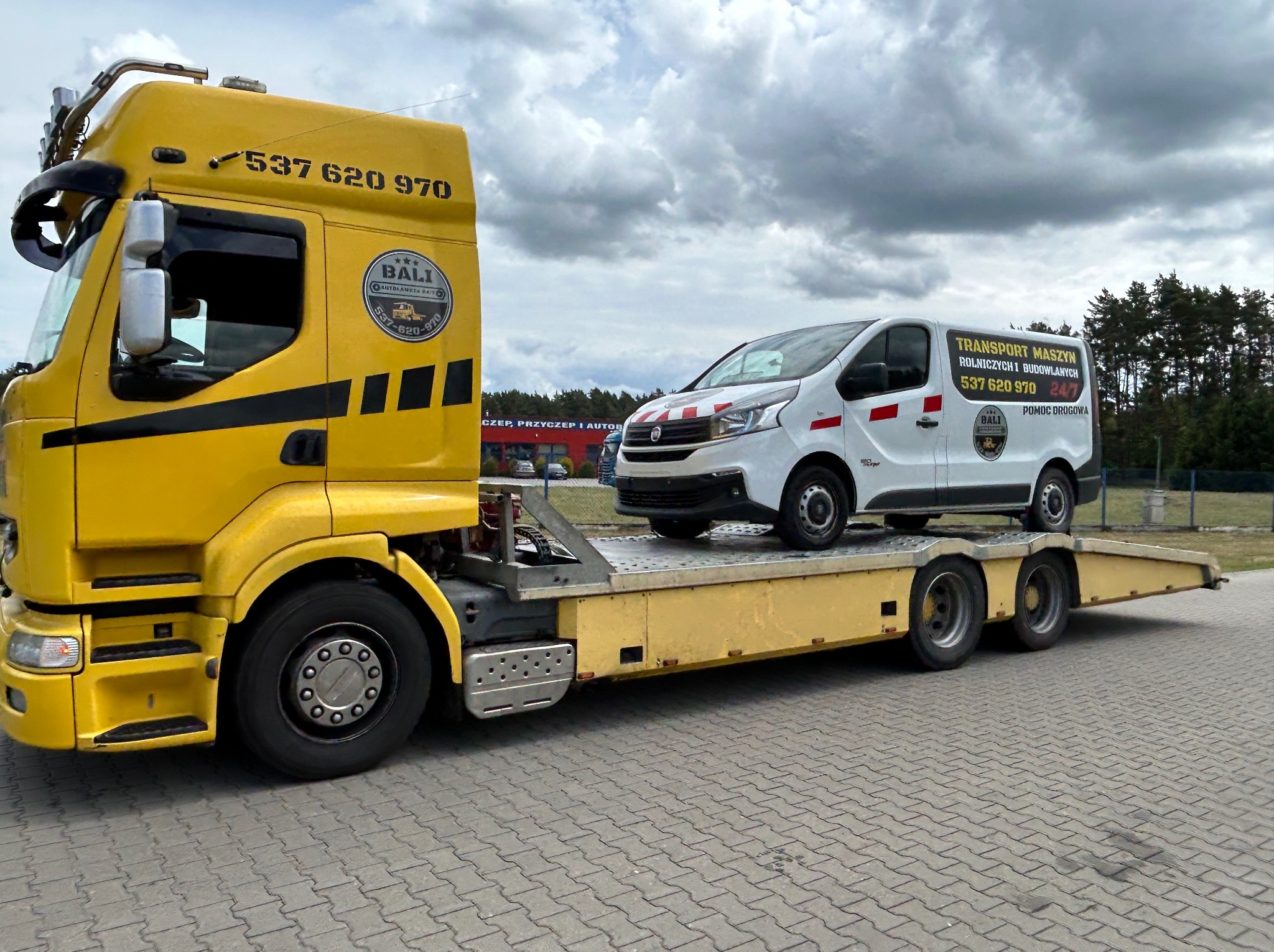 Żółta laweta z białym busem na platformie, na tle pochmurnego nieba. Widoczne logo firmy transportowej i numer telefonu. Pojazd na kostce brukowej.