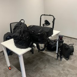 CINEMINDS. - Sprzęt wideo na białym biurku: plecaki, statyw z kamerą, stabilizator. Przygotowania do nagrania w studio z szarą wykładziną.