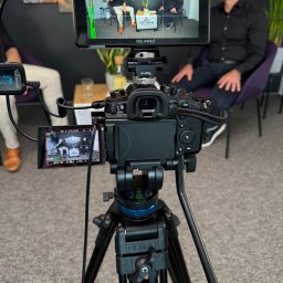 CINEMINDS. - Kamera na statywie nagrywa wywiad z dwoma mężczyznami widocznymi na monitorze. Widoczny mikrofon Saramonic i monitor Feelworld 4K Ultra-Bright.