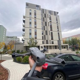 CINEMINDS. - Szary dron DJI Mavic 3 w dłoni, na tle nowoczesnego budynku mieszkalnego i zaparkowanego samochodu. Ujęcie z dołu, pochmurne niebo.