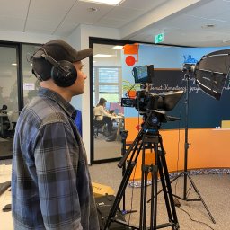 CINEMINDS. - Kamerzysta w trakcie nagrywania lekcji z teleprompterem i oświetleniem studyjnym. Widoczny operator w słuchawkach i kamera na statywie. W tle biuro.