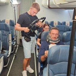 CINEMINDS. - Kamerzysta z profesjonalnym sprzętem filmuje scenę wewnątrz samolotu. Aktor w podróżnej poduszce siedzi na fotelu. Dodatkowe oświetlenie.