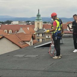 CINEMINDS. - Ekipa na dachu: dekarz z palnikiem i kamerzysta filmujący proces. W tle panorama miasta z kościołem. Praca na wysokościach, zabezpieczenia, profesjonalny sprzęt.